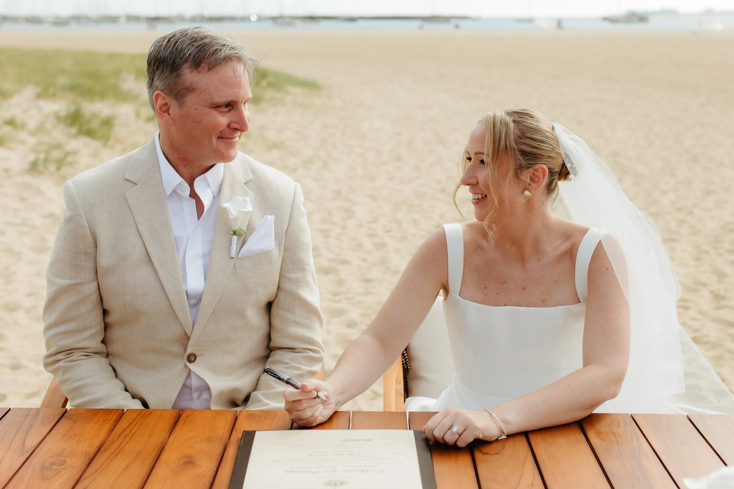 A bride and groom sitting at a wooden table on a beach, with the bride holding a pen and about to sign a document, while the groom looks on.