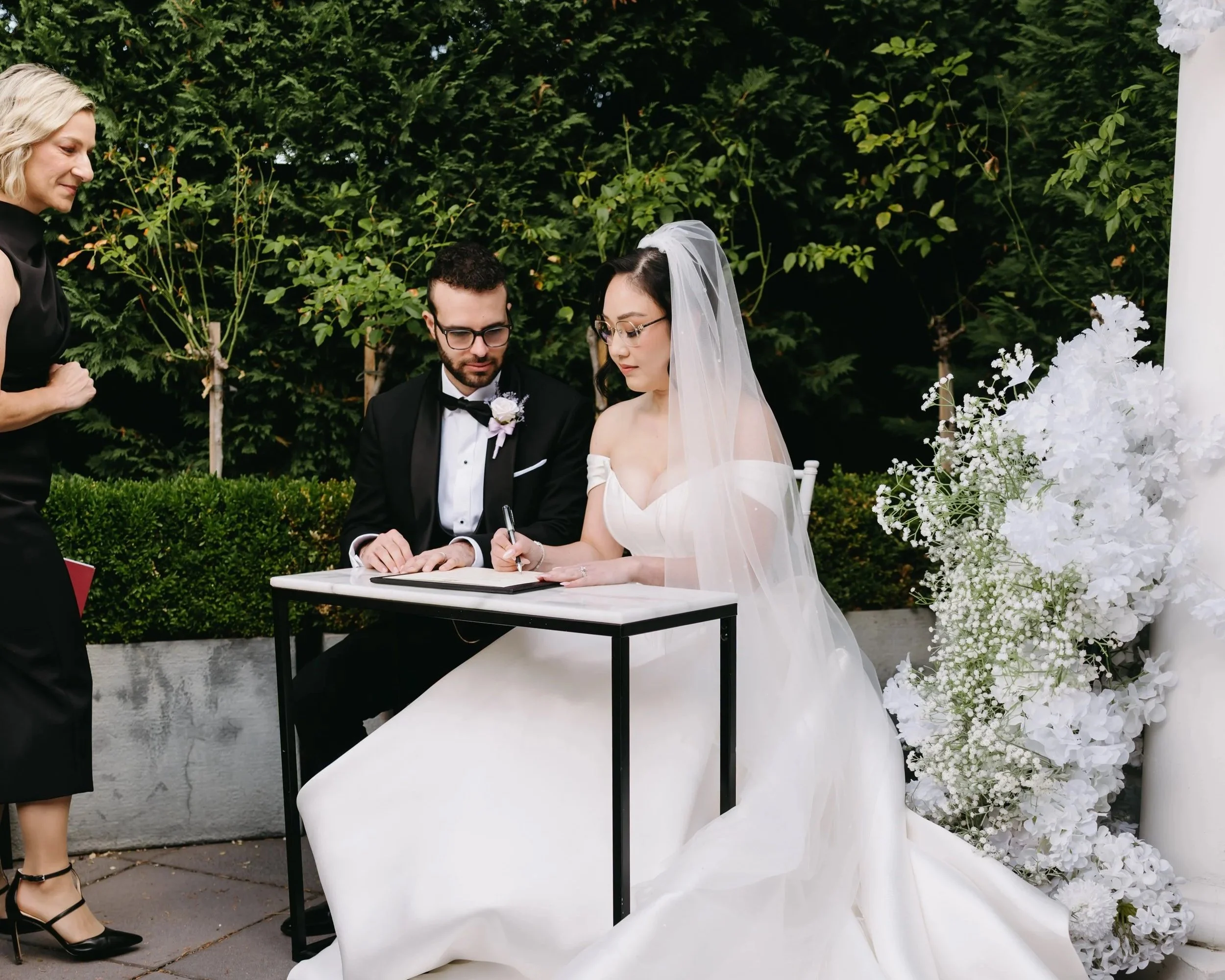 A bride and groom signing a marriage certificate during their wedding ceremony outdoors, with a woman standing nearby and lush greenery in the background.