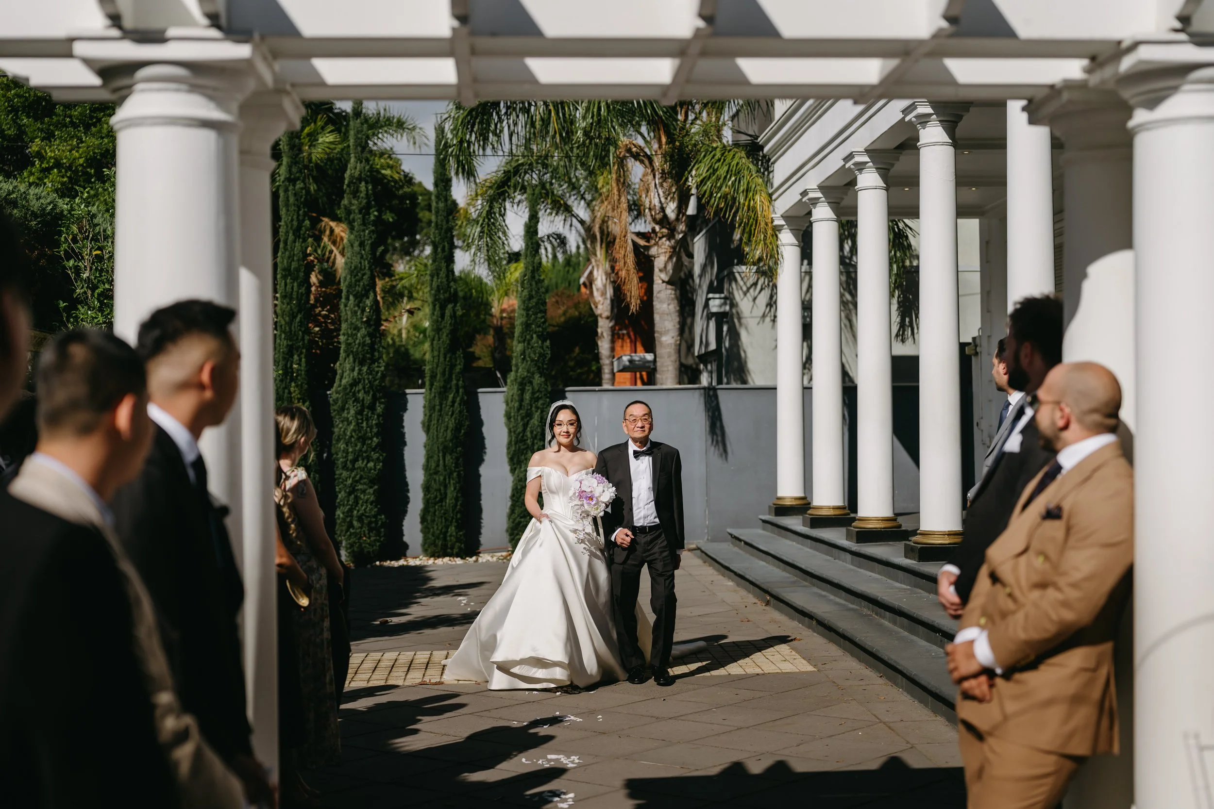 A bride walking down the aisle with her father during a wedding ceremony outdoors. The bride is wearing a white wedding dress and holding a bouquet, while guests are standing on either side of the aisle under white columns.