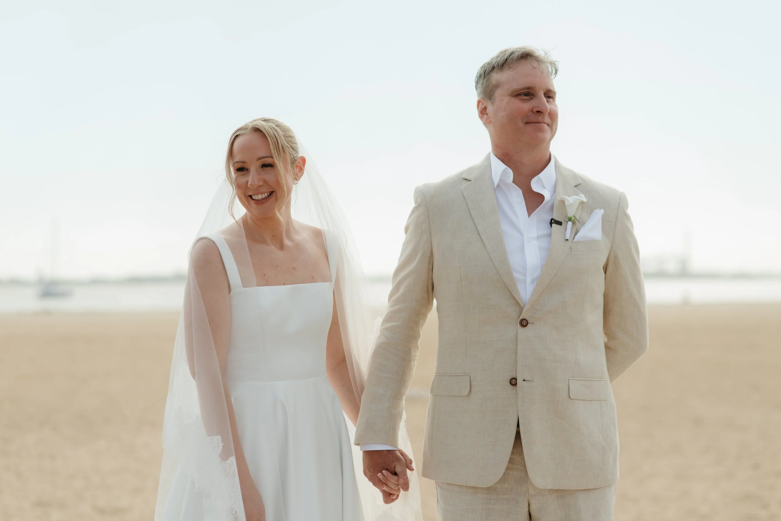 A bride and groom holding hands on a beach, smiling, dressed in wedding attire with the bride in a white gown and veil, and the groom in a beige suit with a white shirt and boutonniere, overcast sky in the background.