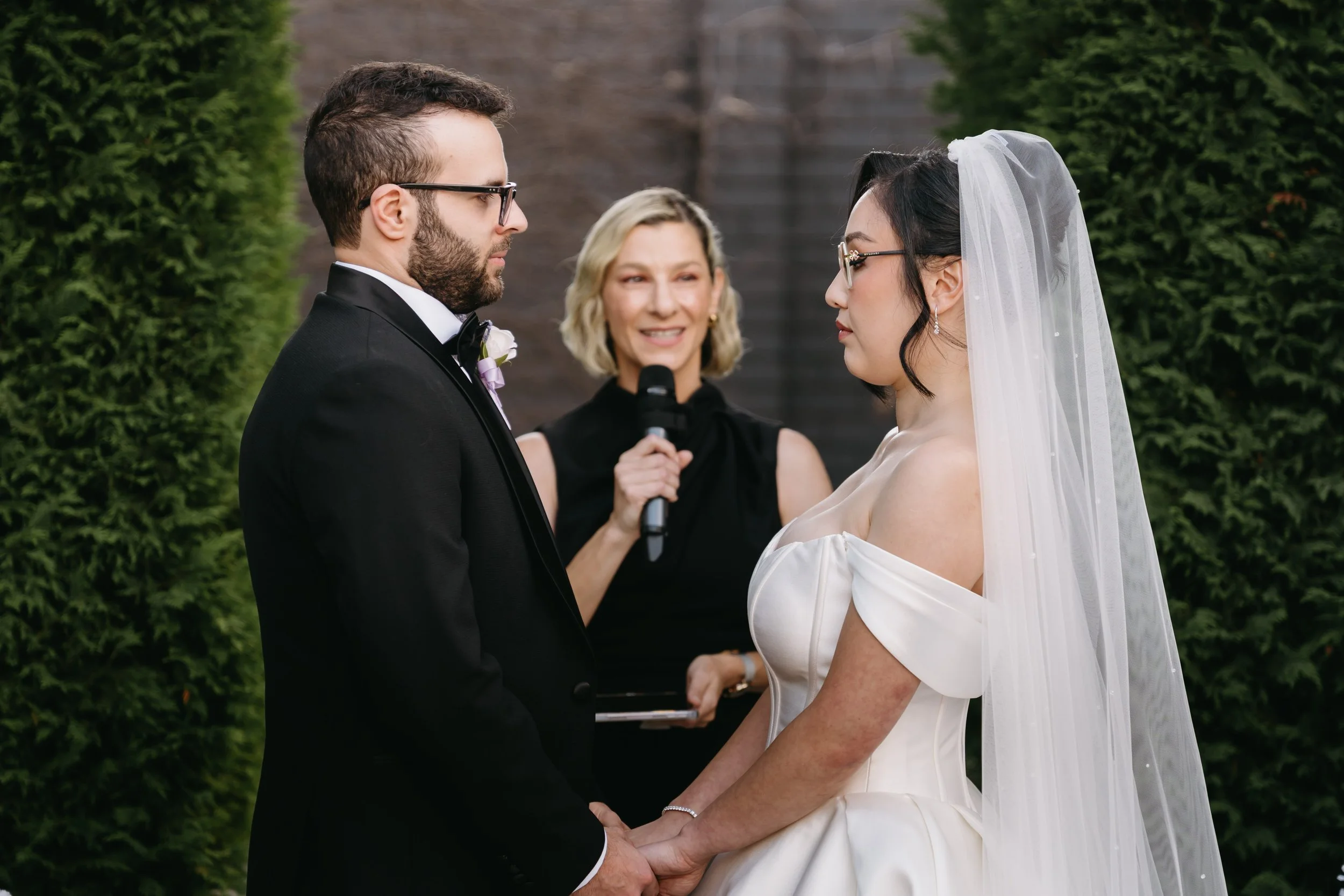 A wedding ceremony with a bride and groom holding hands and facing each other, with an officiant in the background holding a microphone outdoors, surrounded by green trees.