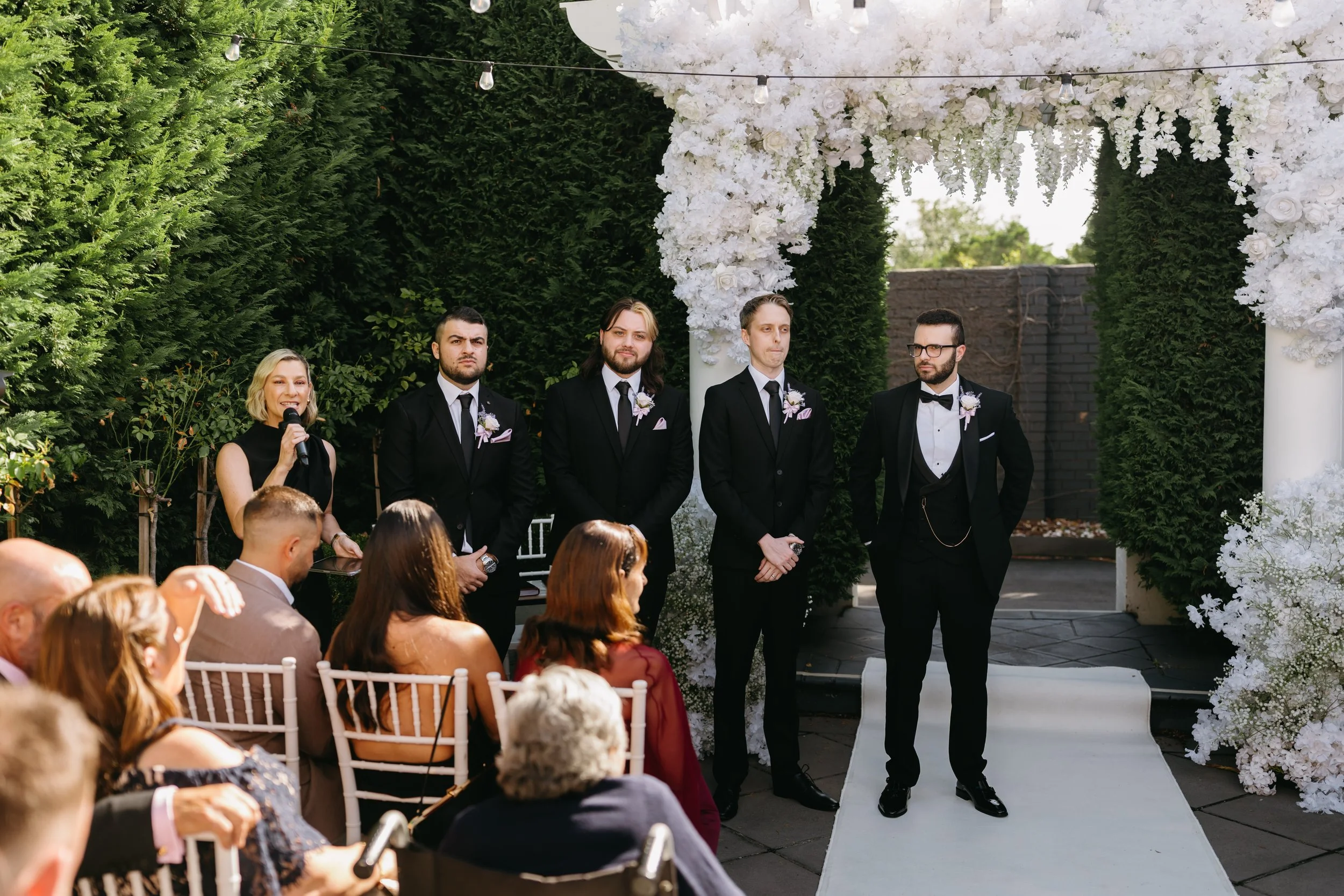 A wedding ceremony taking place outdoors with guests seated and a decorated floral arch under string lights.