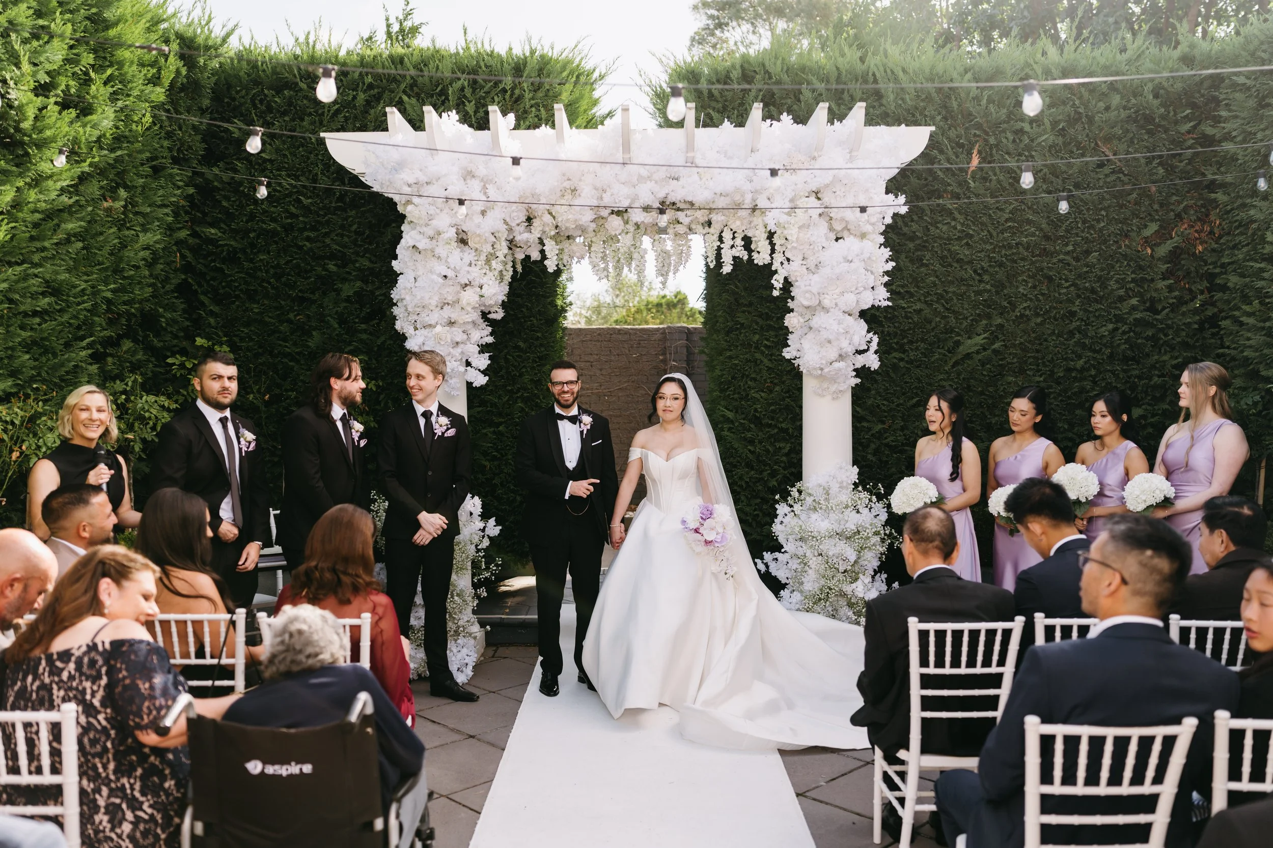 A wedding ceremony outdoors with a bride and groom standing under a floral arch, surrounded by bridesmaids and groomsmen, with guests seated around them.