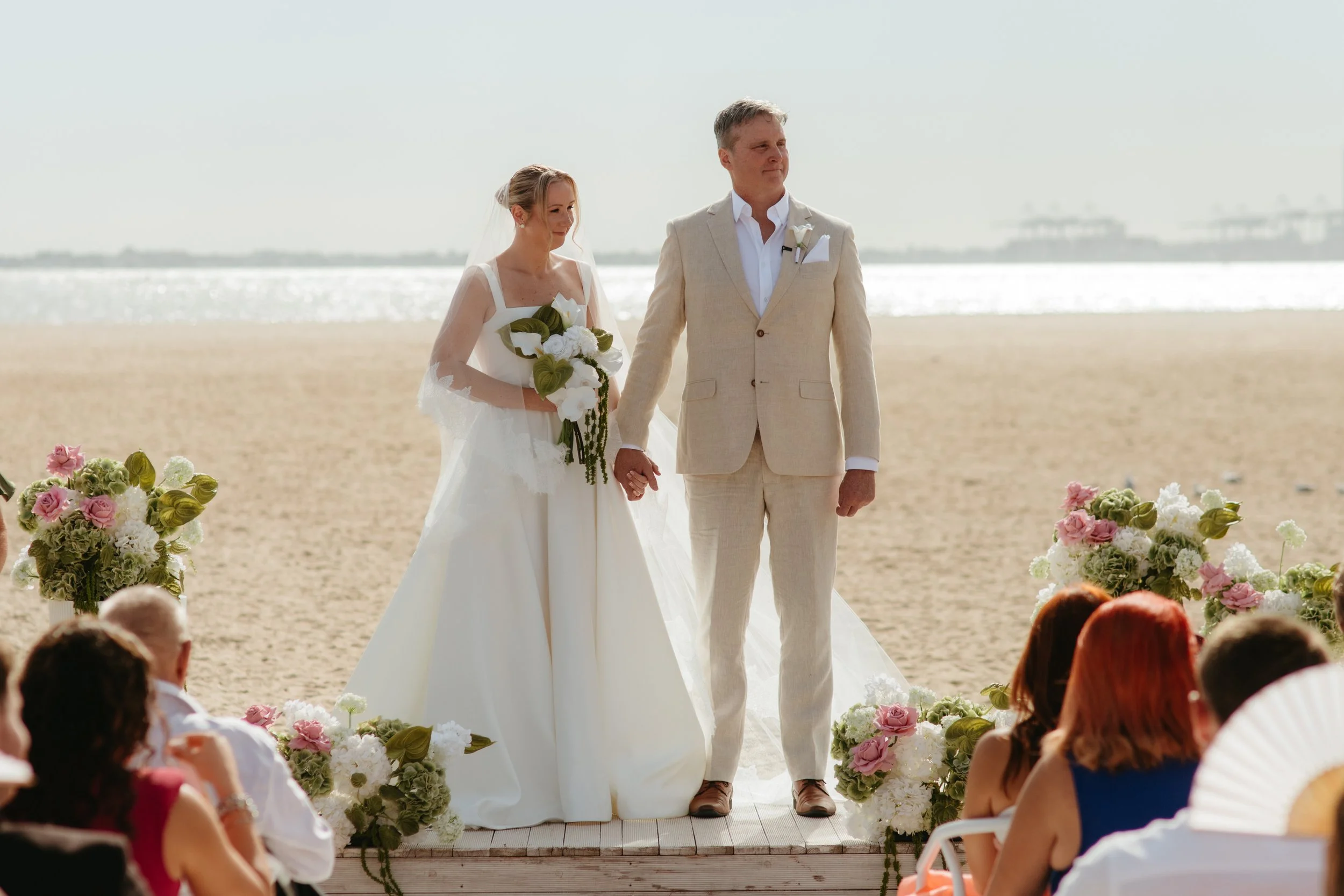 A bride and groom holding hands during their beach wedding ceremony, surrounded by flowers and guests seated in front of them, with the ocean and distant bridge in the background.