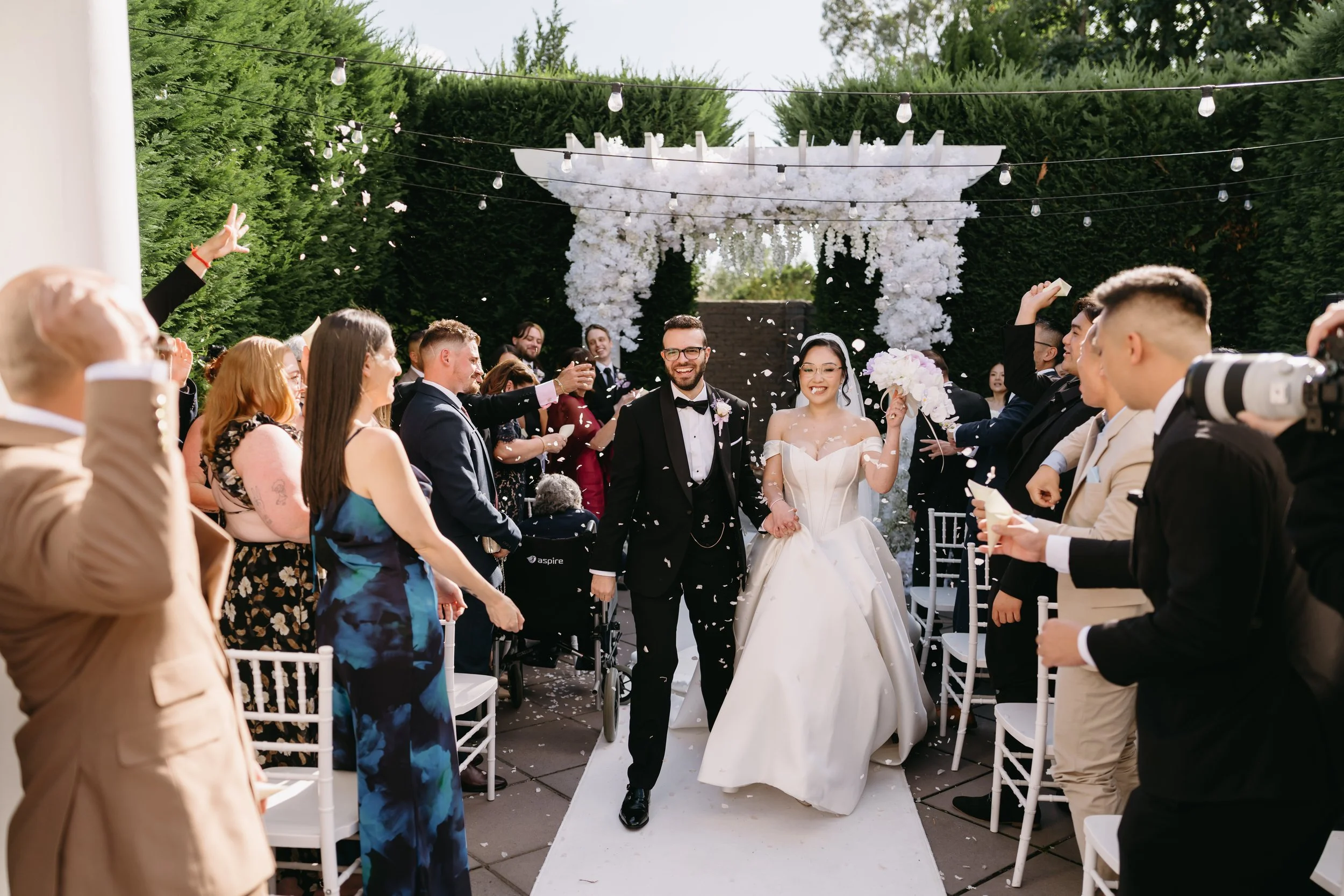 A wedding ceremony outdoors with a bride and groom walking down the aisle, surrounded by guests. The bride is wearing a white wedding gown and holding a bouquet, and the groom is in a tuxedo. The ceremony area is decorated with white flowers and stri