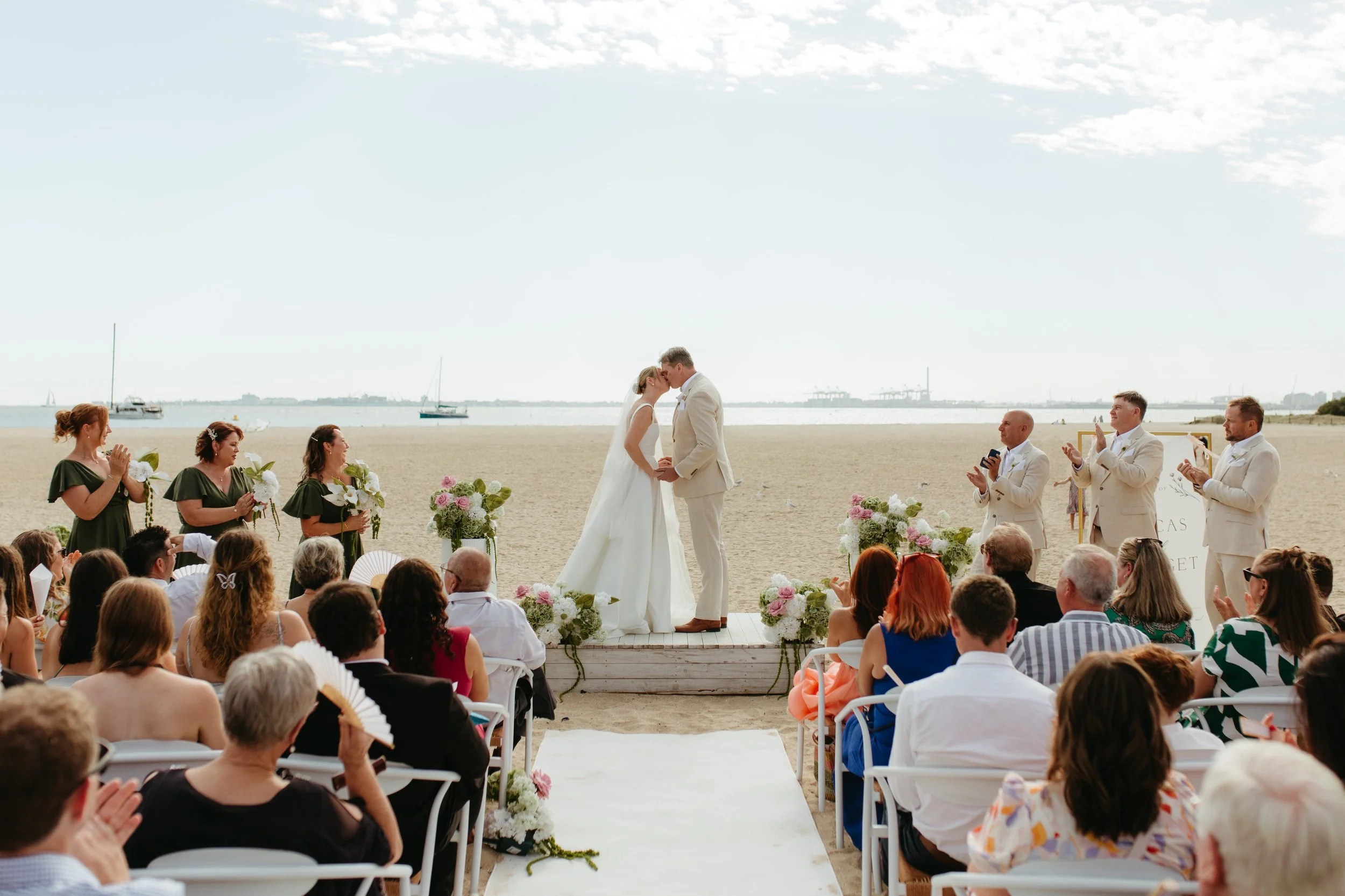 A beach wedding ceremony with a bride and groom kissing, surrounded by bridesmaids and groomsmen, with guests seated in front on chairs, ocean and sailboats in the background under a partly cloudy sky.