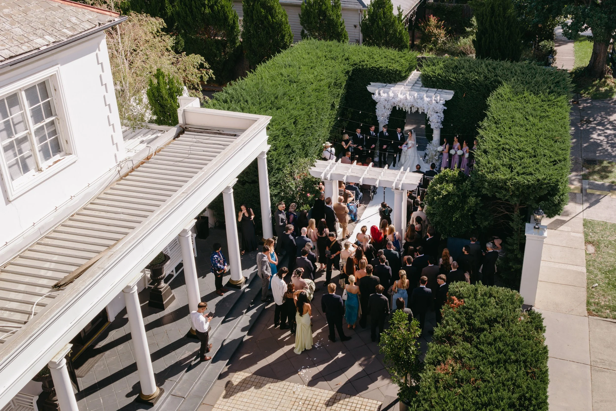 A wedding ceremony taking place outdoors in a lush garden area with guests gathered. The bride and groom stand under a floral arch with a small choir or group of singers nearby. There are many guests dressed formally, and the setting is a sunny day w