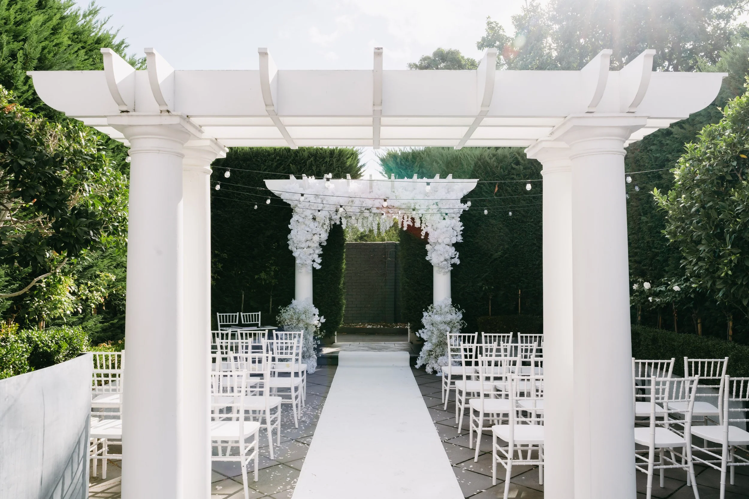 An outdoor wedding setup with white chairs arranged on either side of a white aisle runner, white floral decorations at the entrance of an archway, and string lights hanging above, surrounded by green trees.