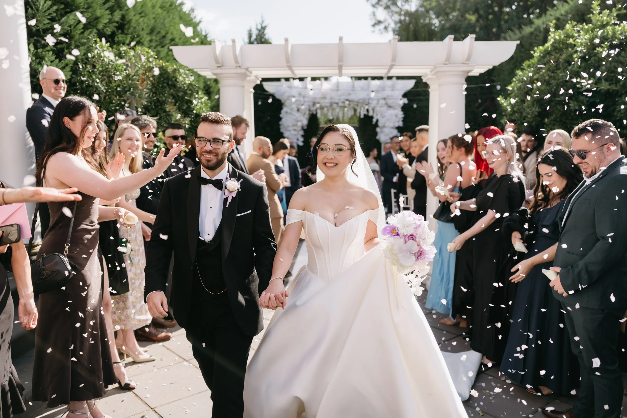A bride and groom walking hand in hand through a crowd of guests who are throwing flower petals at a wedding ceremony outside under a white pergola. The bride is wearing a white strapless wedding gown and glasses, while the groom is dressed in a blac