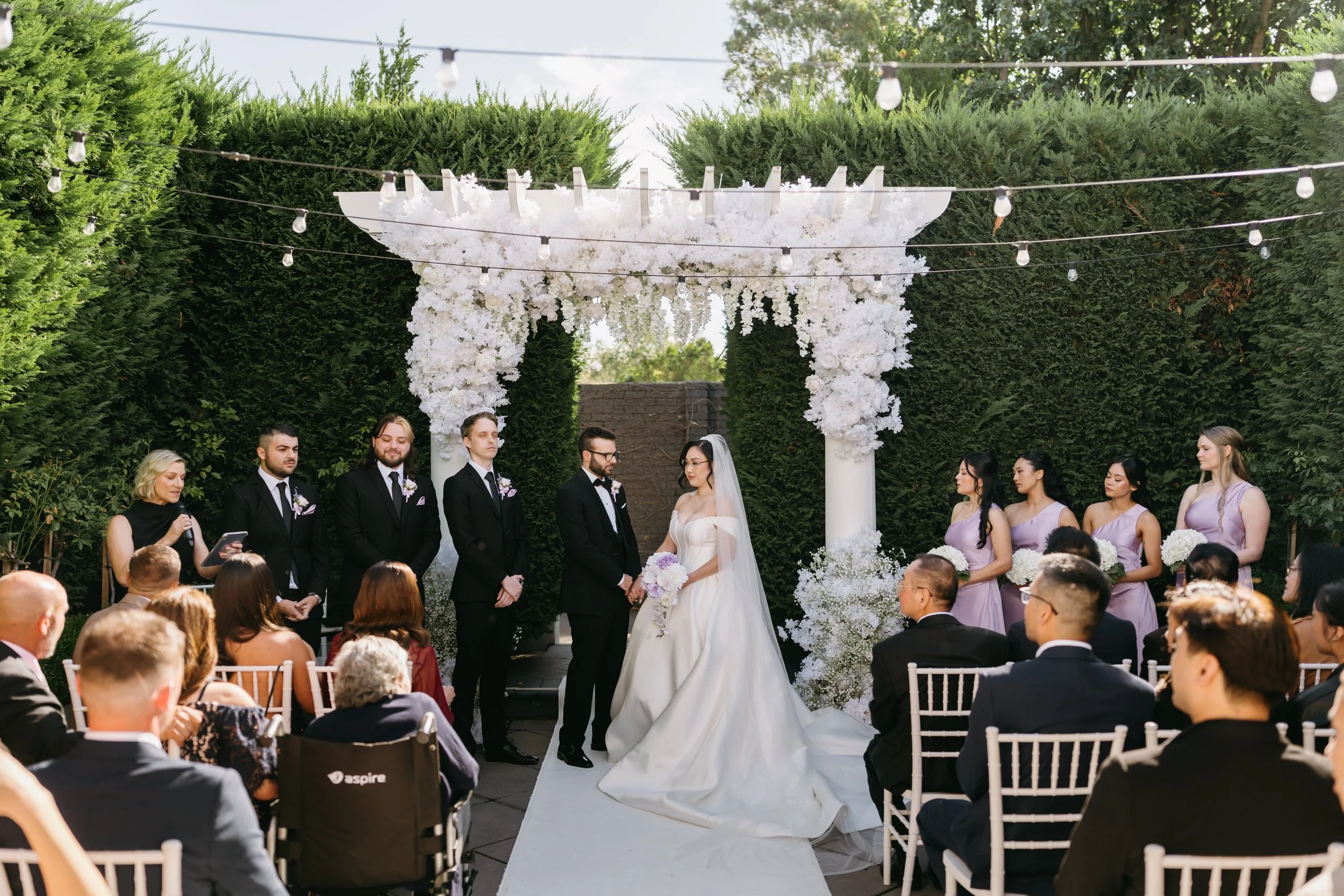 A wedding ceremony takes place outdoors under a decorative white floral arch with greenery behind. The bride and groom stand facing each other, with the bride holding a bouquet. Bridesmaids in lavender dresses and groomsmen in black tuxedos with purp