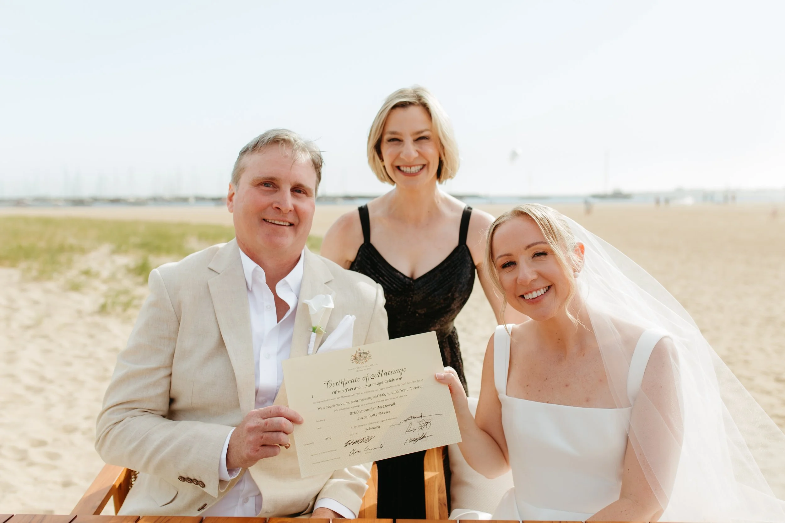 A group of three people with light skin on a beach, celebrating a wedding. The man in a light suit holds a marriage certificate, and the women, one in a black dress and the other in a white wedding dress, smile happily.