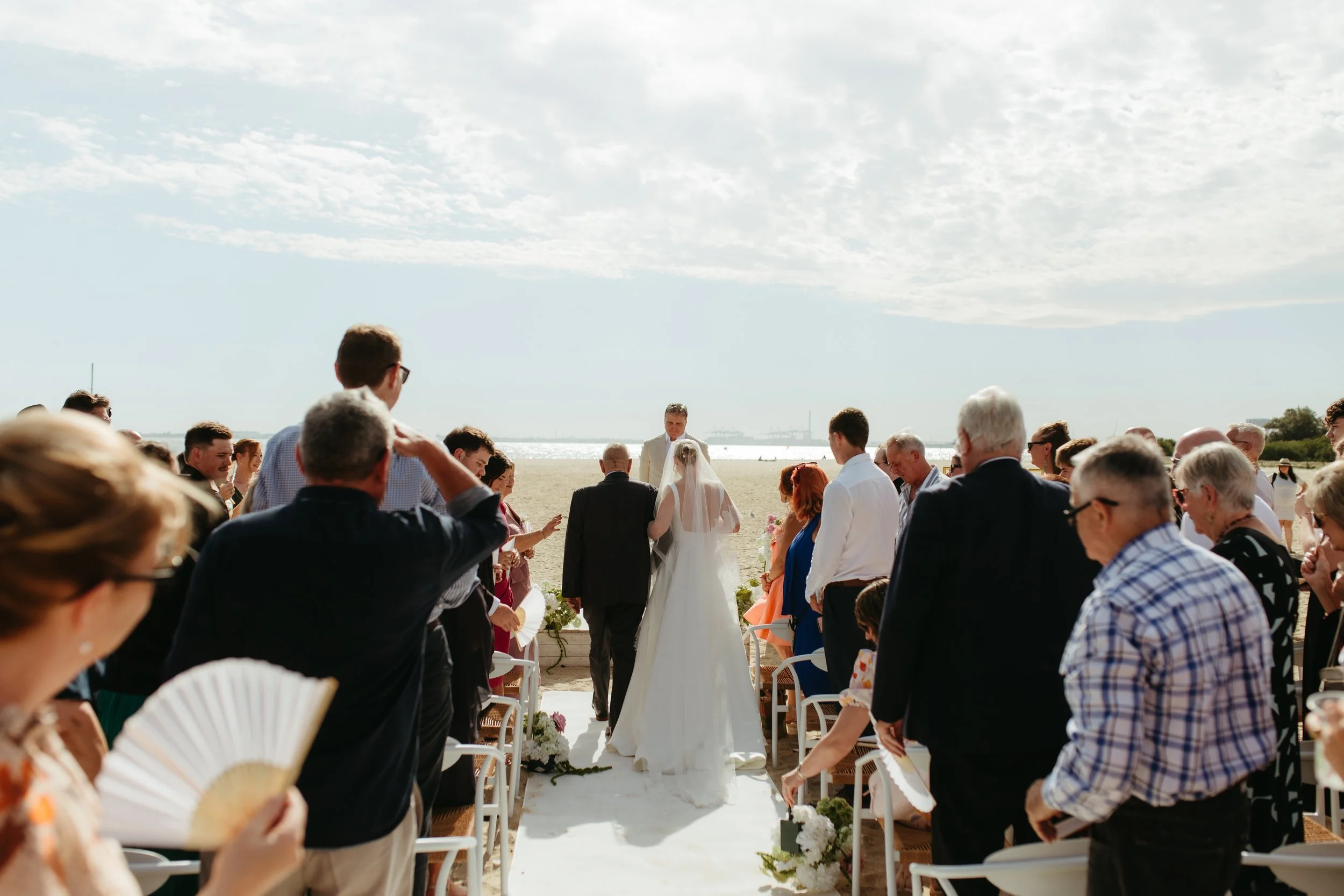 A wedding ceremony taking place on a beach with people gathered around a bride and groom, the bride wearing a white dress and veil and the groom in a suit, under a cloudy sky.