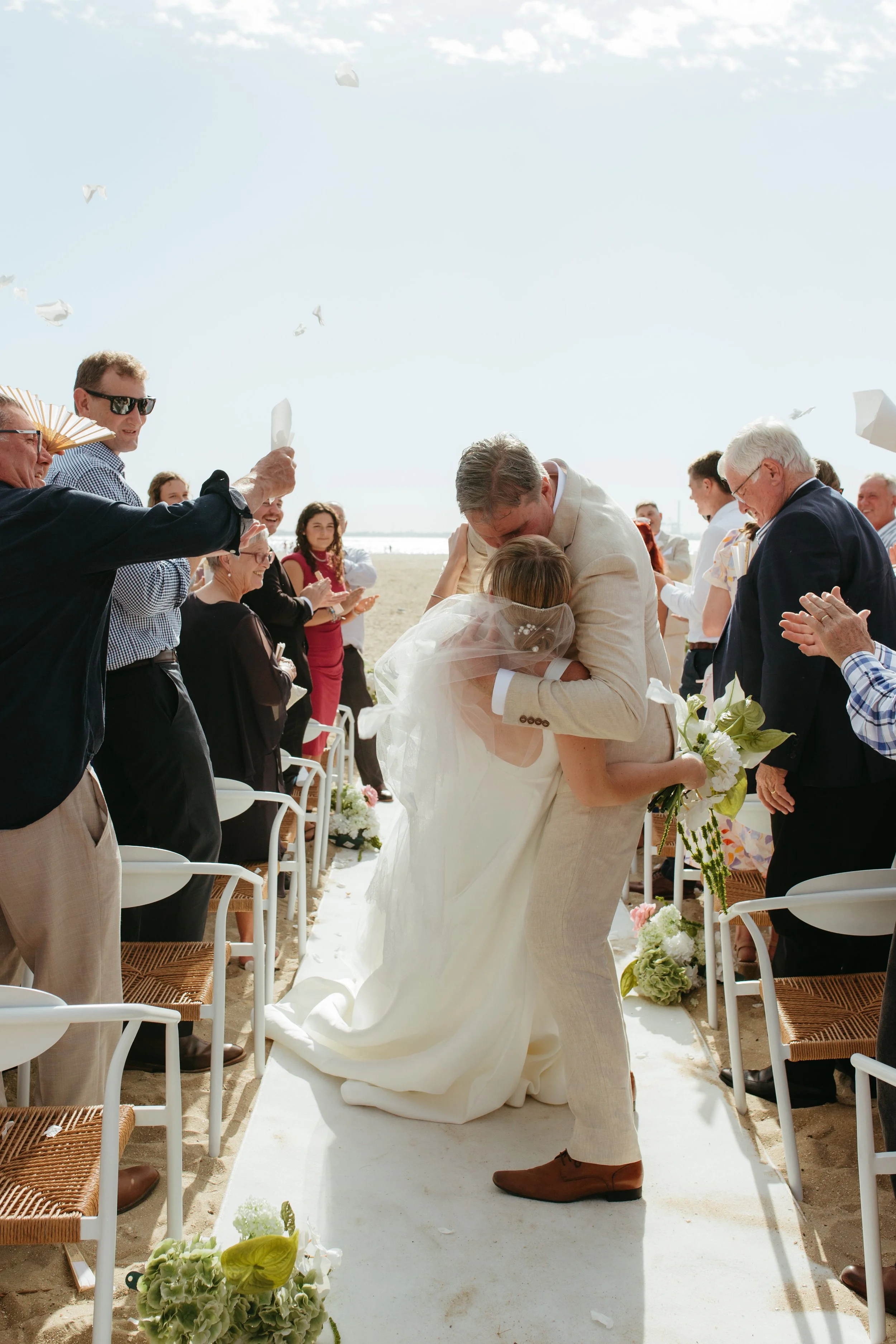 A wedding ceremony takes place outdoors on a beach with a white aisle runner, floral arrangements, and seated guests. The bride and groom are embracing in front of friends and family.