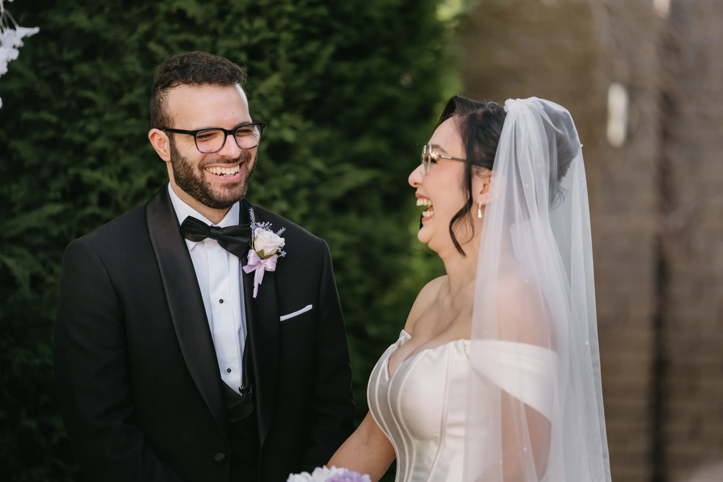 A bride and groom with glasses smiling at each other outdoors during a wedding ceremony.