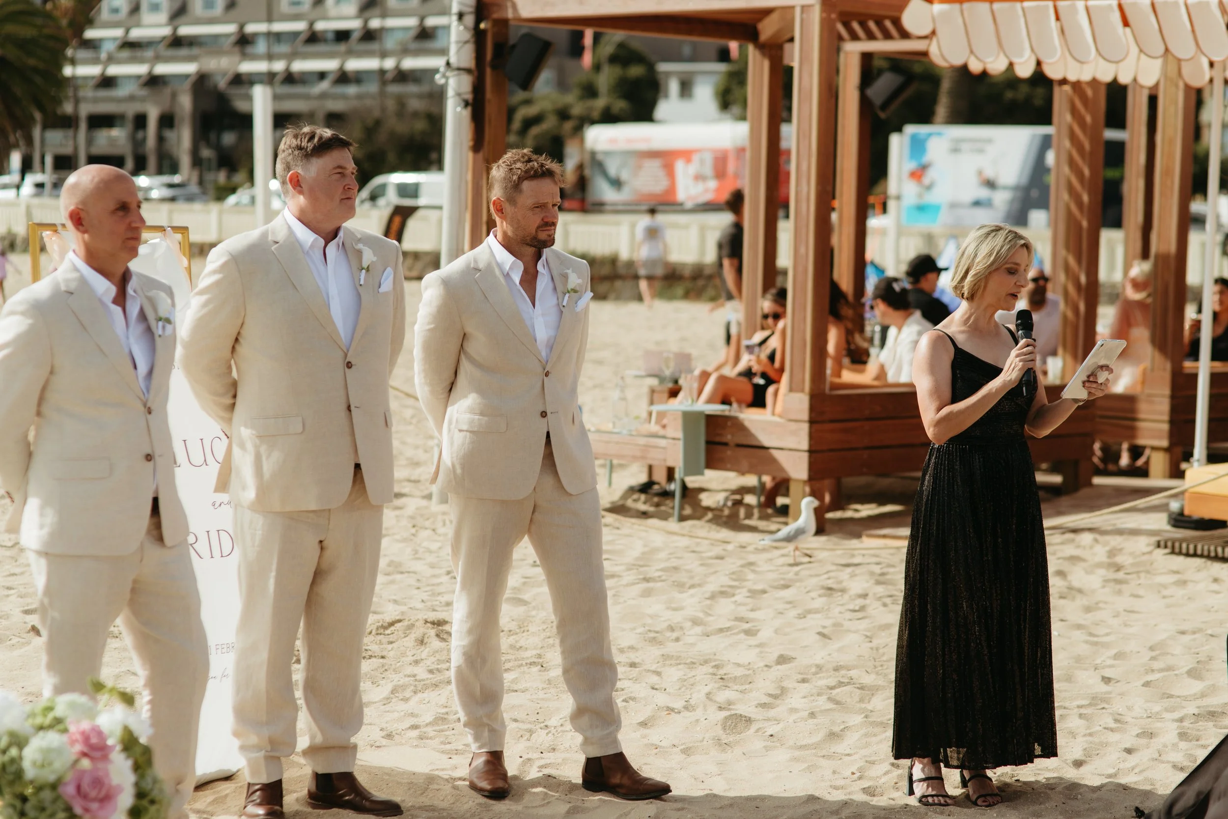Three men dressed in cream suits standing on a sandy beach during a wedding ceremony, with a woman reading from a tablet or phone nearby. There are people seated in the background and structures with wooden frames and striped awnings.