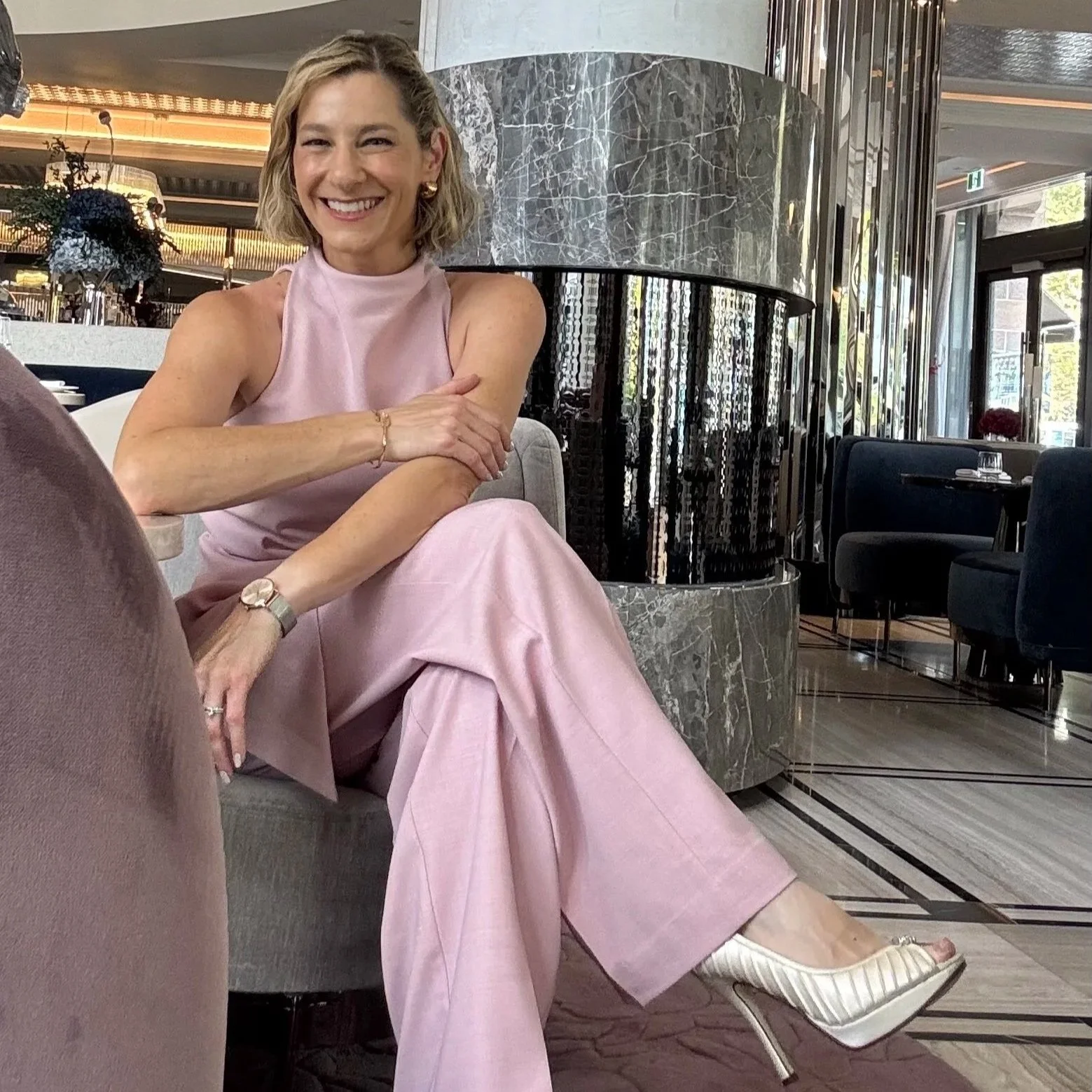 Marriage Celebrant Olivia Ferraro. A woman in a pink sleeveless top and matching pink pants, sitting comfortably in a modern restaurant or hotel lobby with marble, mirrored, and glass decor, smiling at the camera. The Royce Hotel, Melbourne