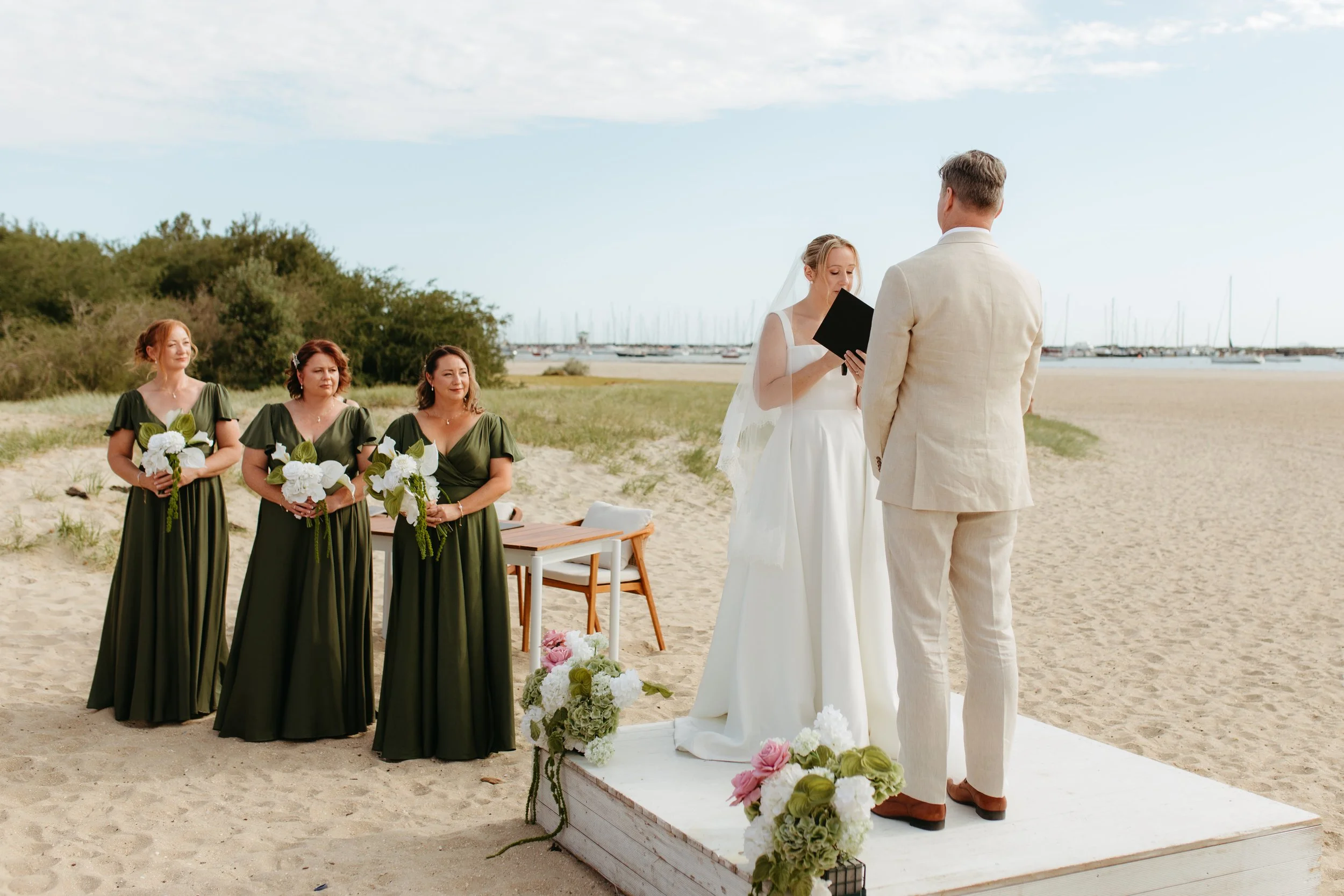 A beach wedding ceremony with a bride and groom exchanging vows, three bridesmaids holding bouquets, and a floral arrangement on a wooden platform, with boats docked in the background.