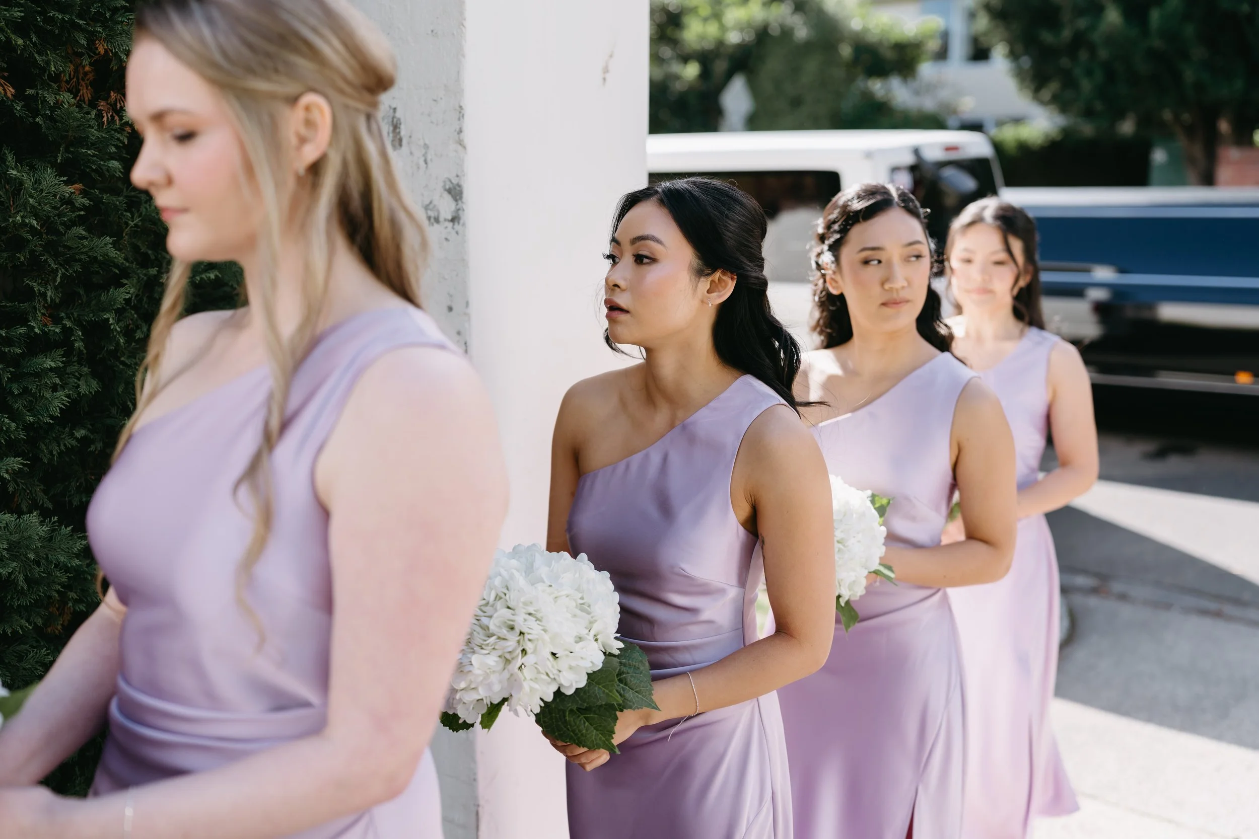 A line of women in lavender dresses holding white bouquets, standing outdoors in sunlight, with a white wall and greenery in the background.