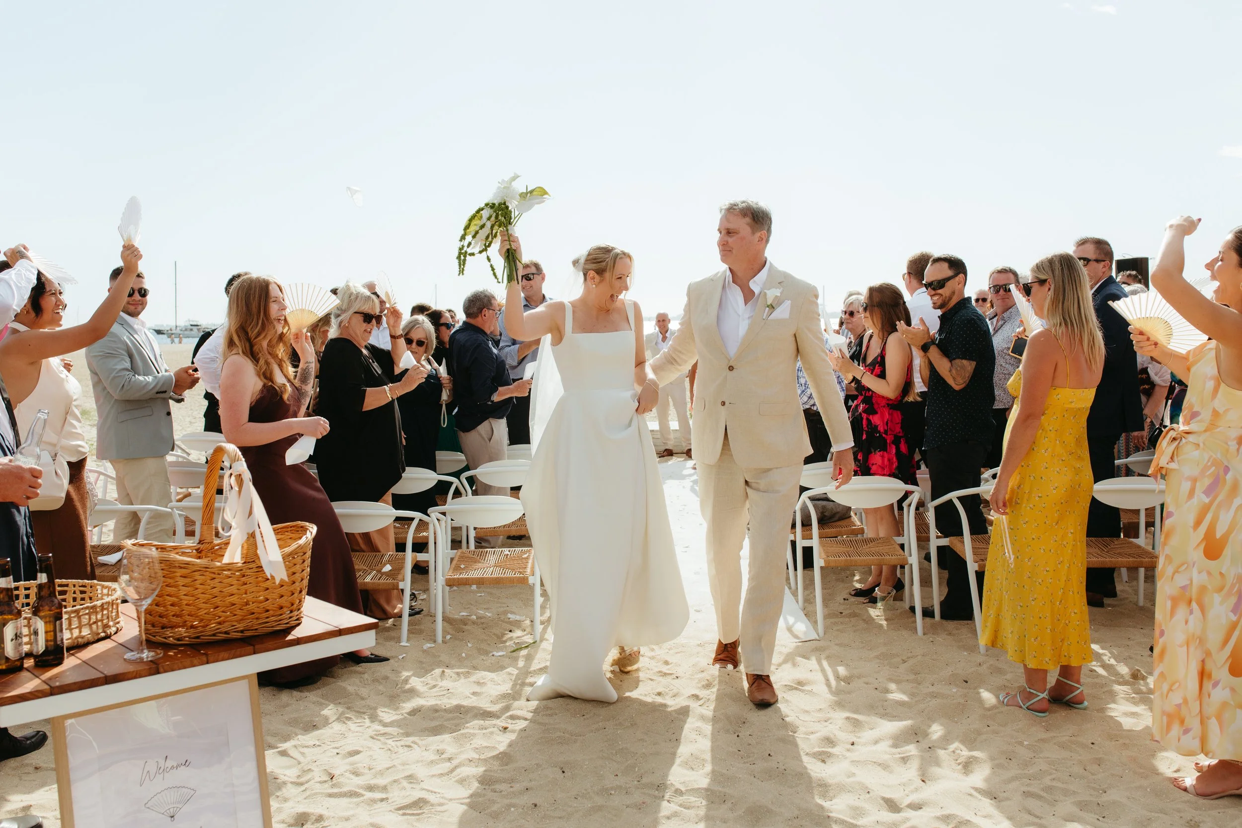 A bride and groom walking on a sandy beach during their wedding ceremony, surrounded by guests holding fans and standing under sunny skies.