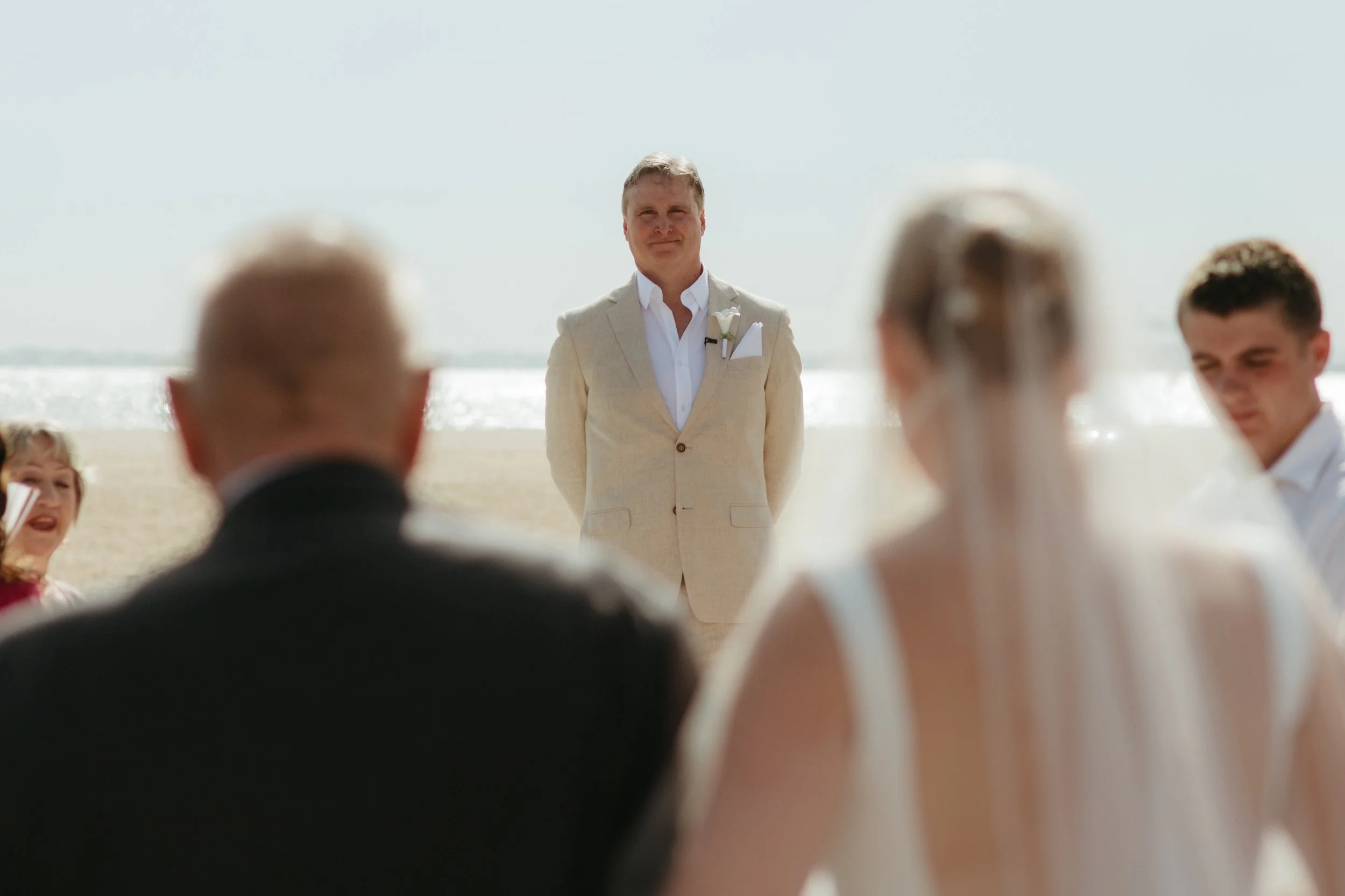 A wedding ceremony on the beach with a groom in a beige suit and a white shirt standing in front of guests. Beach and ocean in the background.