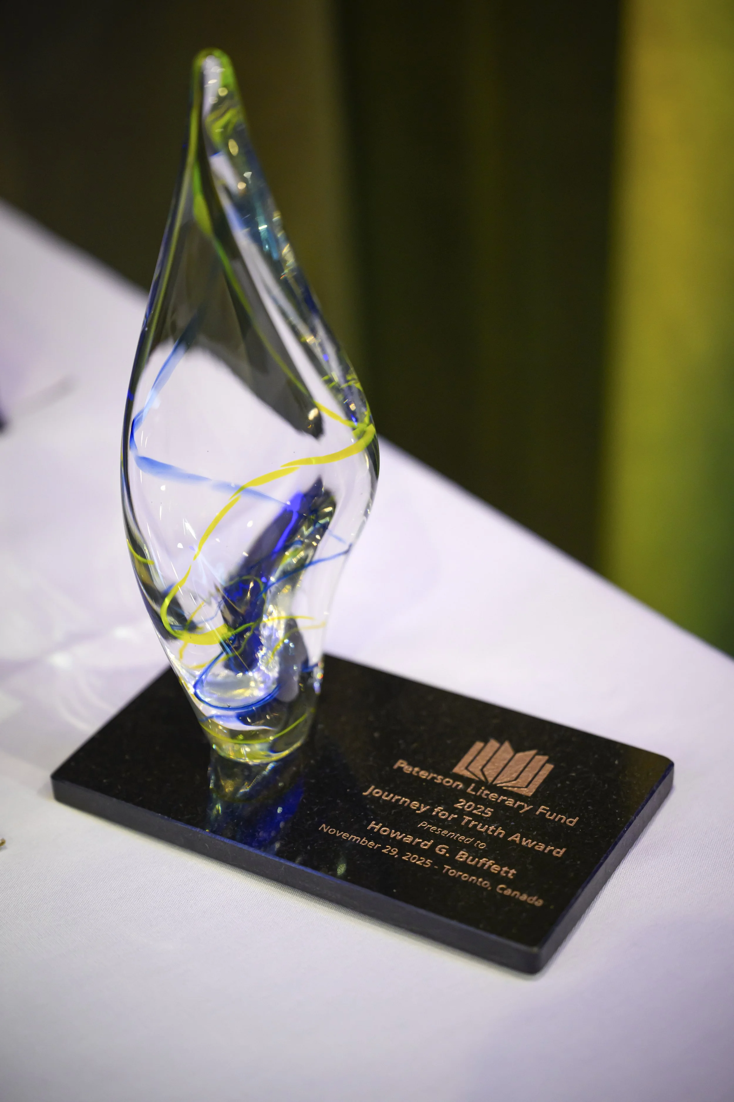 A glass trophy with colorful swirling patterns, mounted on a black base with engraved text and logos, displayed on a white surface with a dark background.
