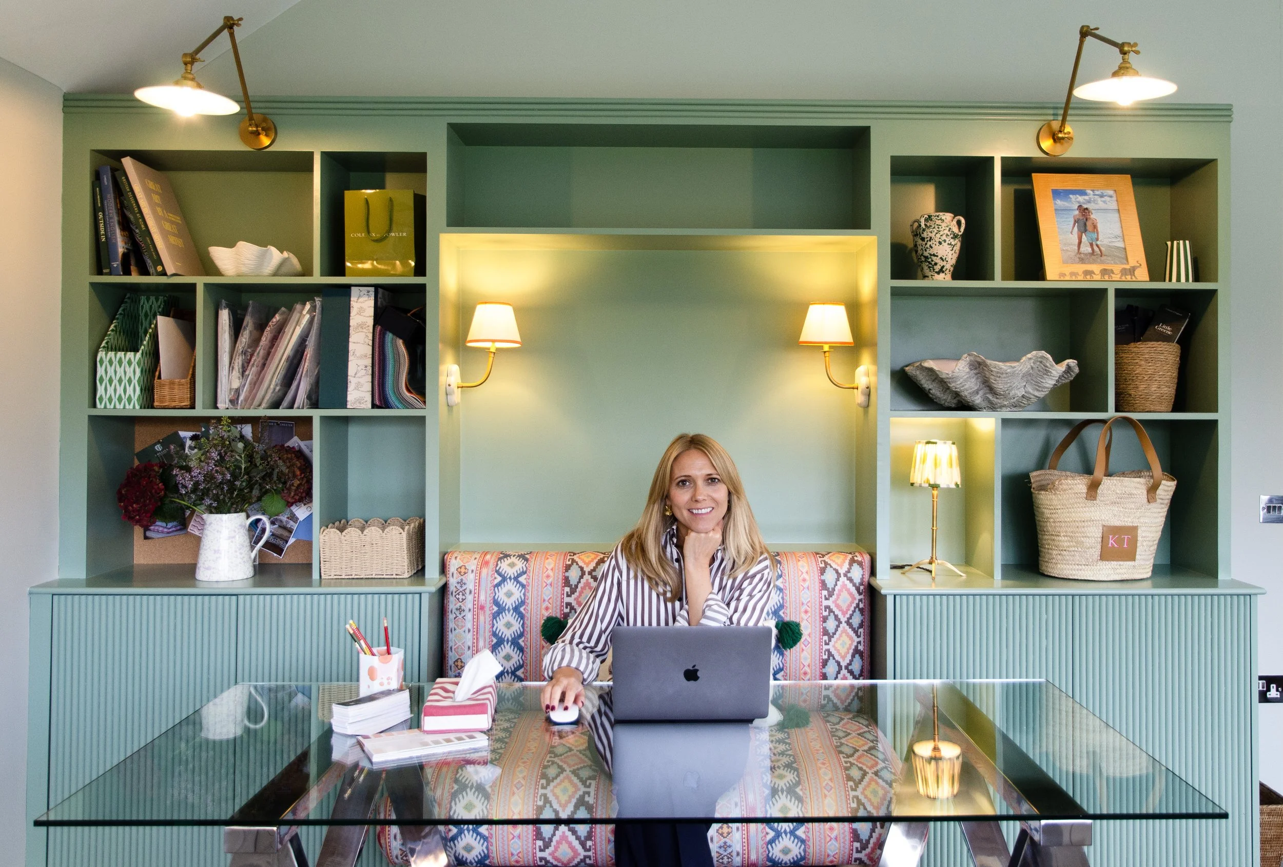 A woman sitting at a glass-topped desk with a laptop in a room with green walls and built-in shelving, illuminated by wall-mounted and table lamps, decorated with books, framed photograph, vase, and decorative items.