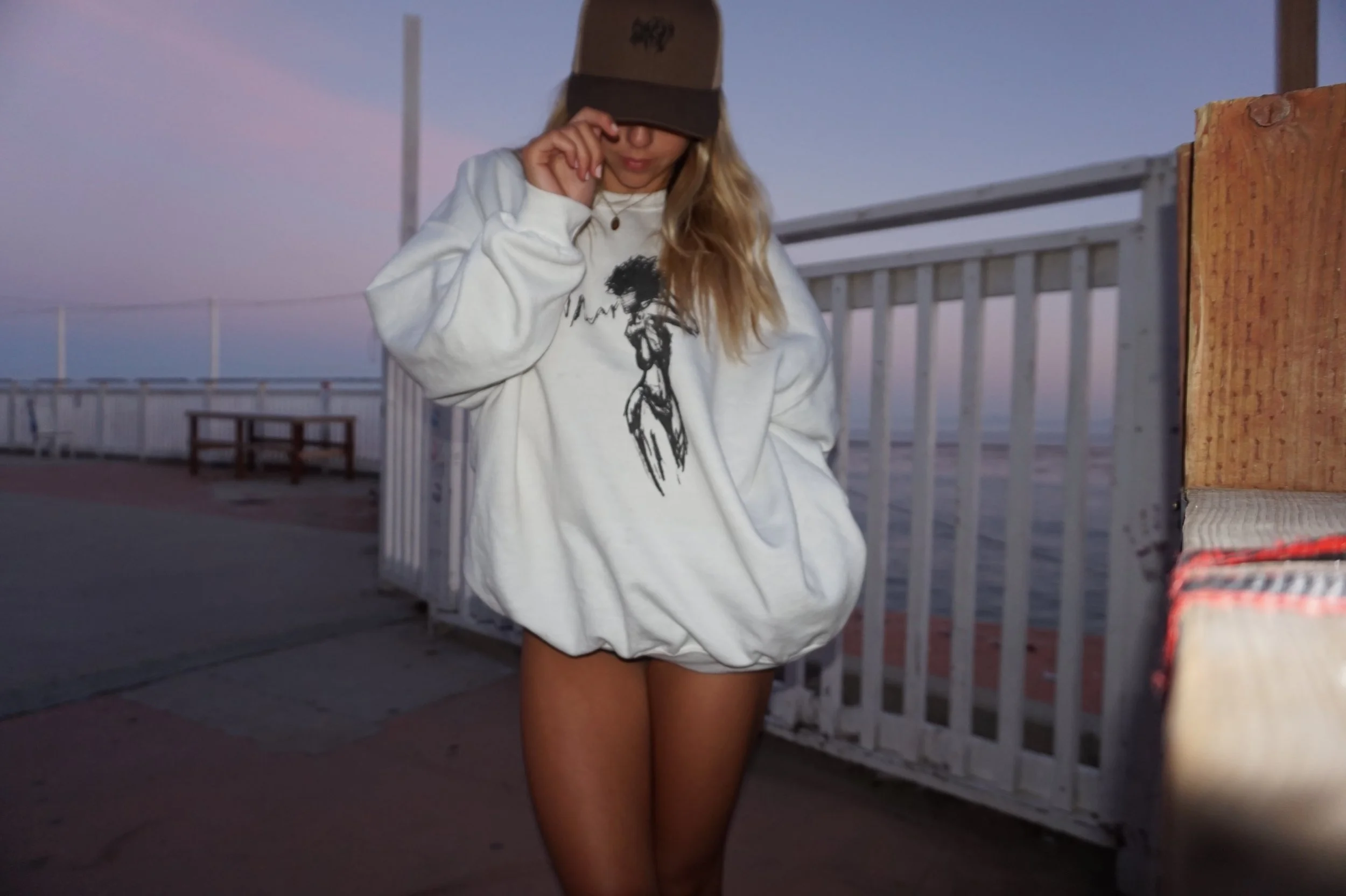 Person in oversized sweatshirt and baseball cap standing on boardwalk at dusk.