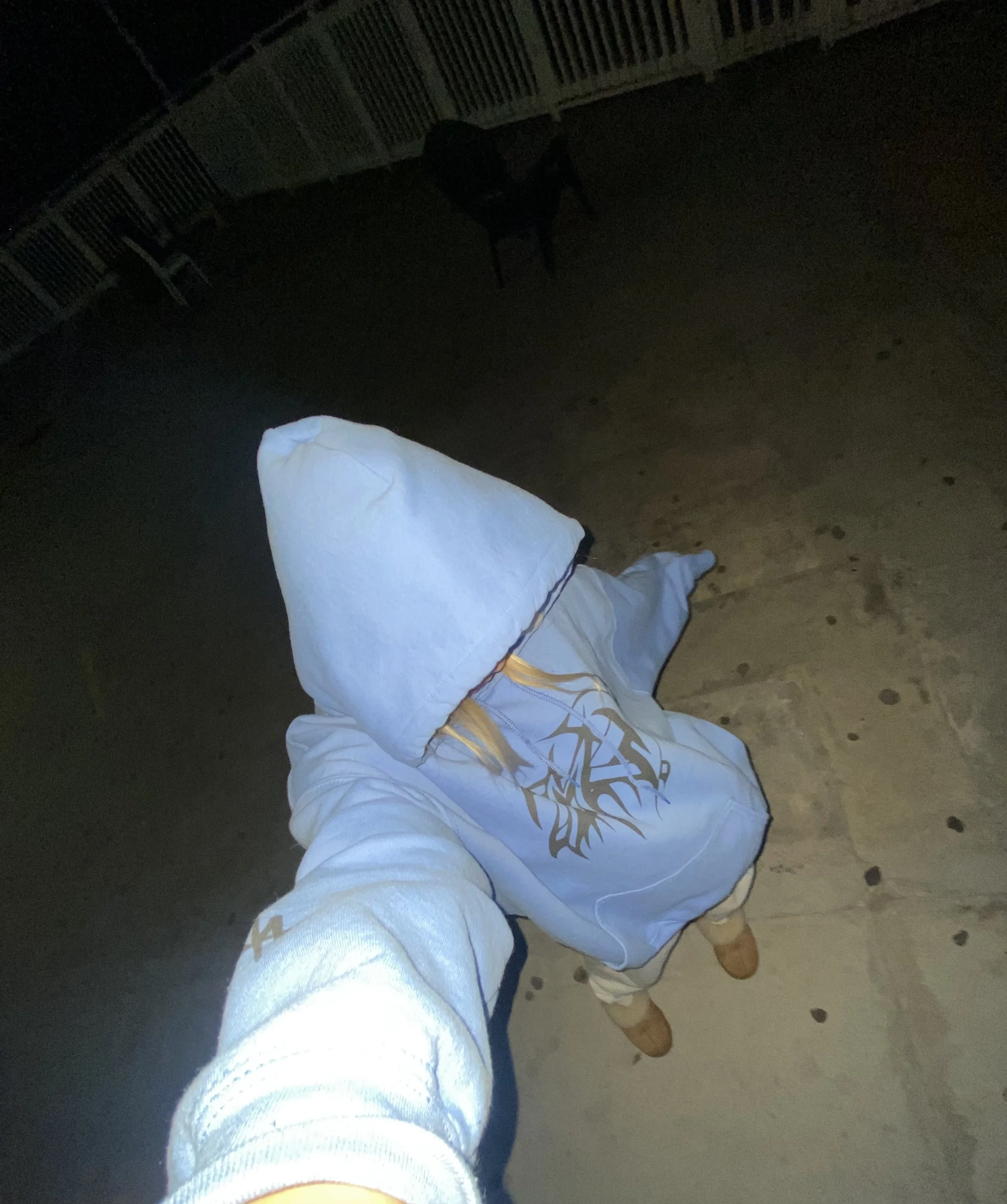 Person wearing a blue hoodie with a graphic design, standing outdoors on a patio at night, with a white fence and chairs in the background.