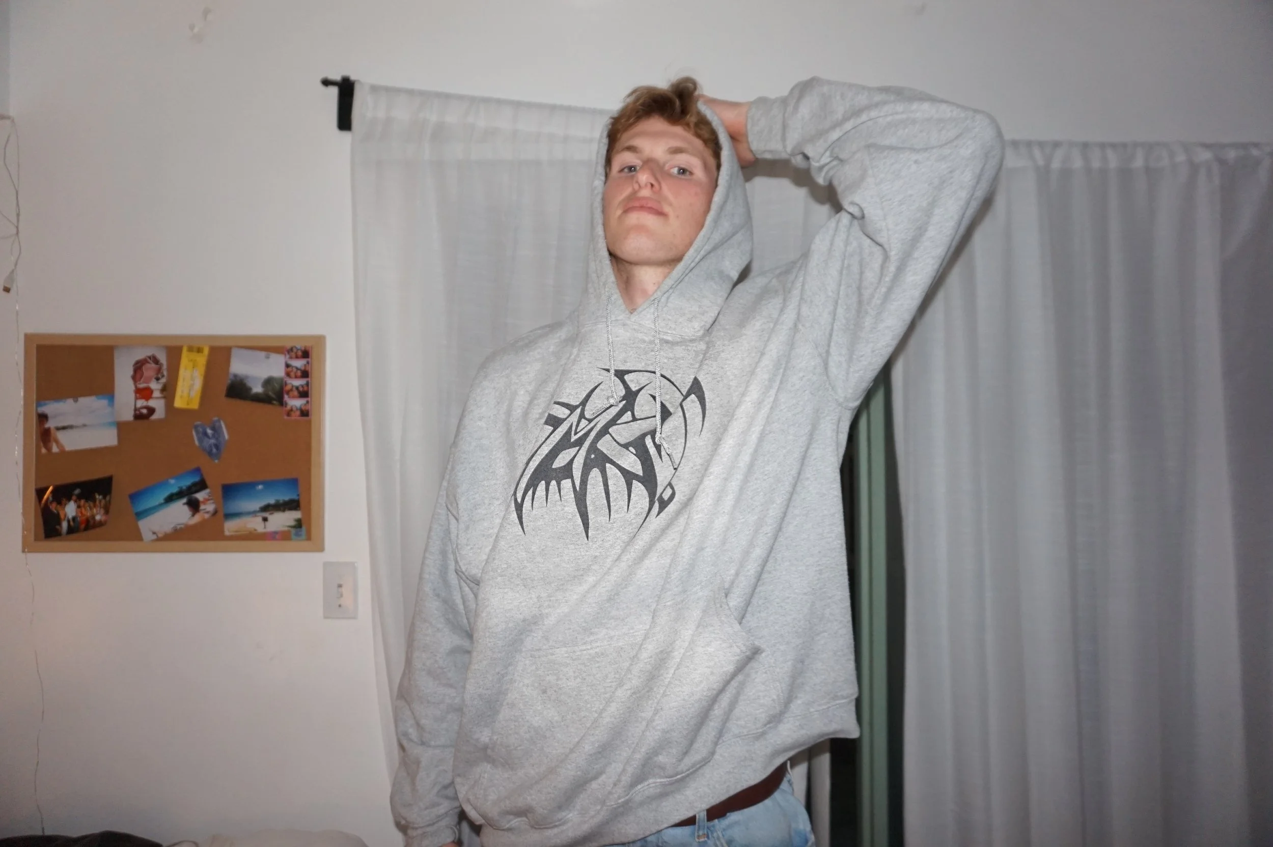 Person in a gray hoodie standing in a room with a cork board and curtains in the background.