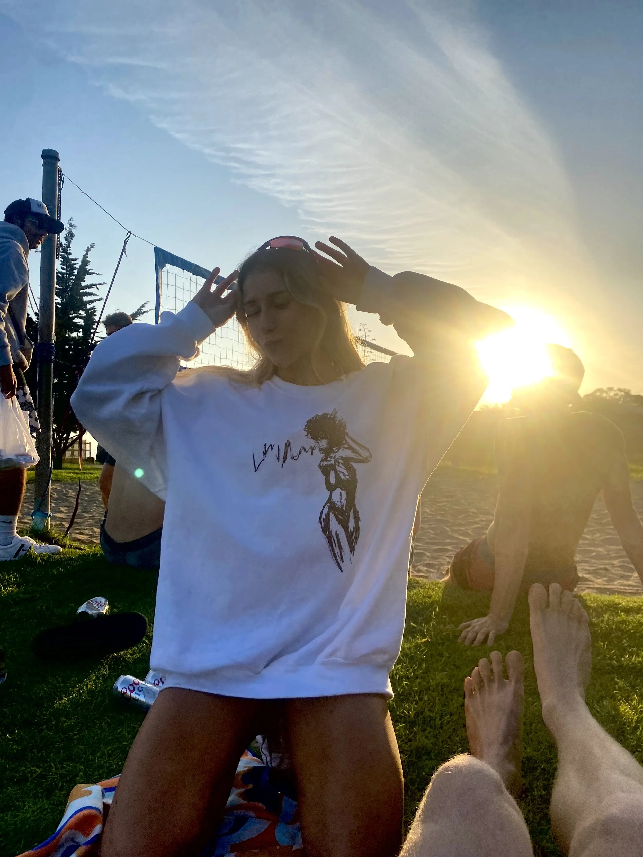 A person in a white sweatshirt with a graphic design stands outdoors at sunset. They are surrounded by people sitting on the grass near a volleyball net, with the sun shining brightly in the background. Visible are some feet in the foreground and a b