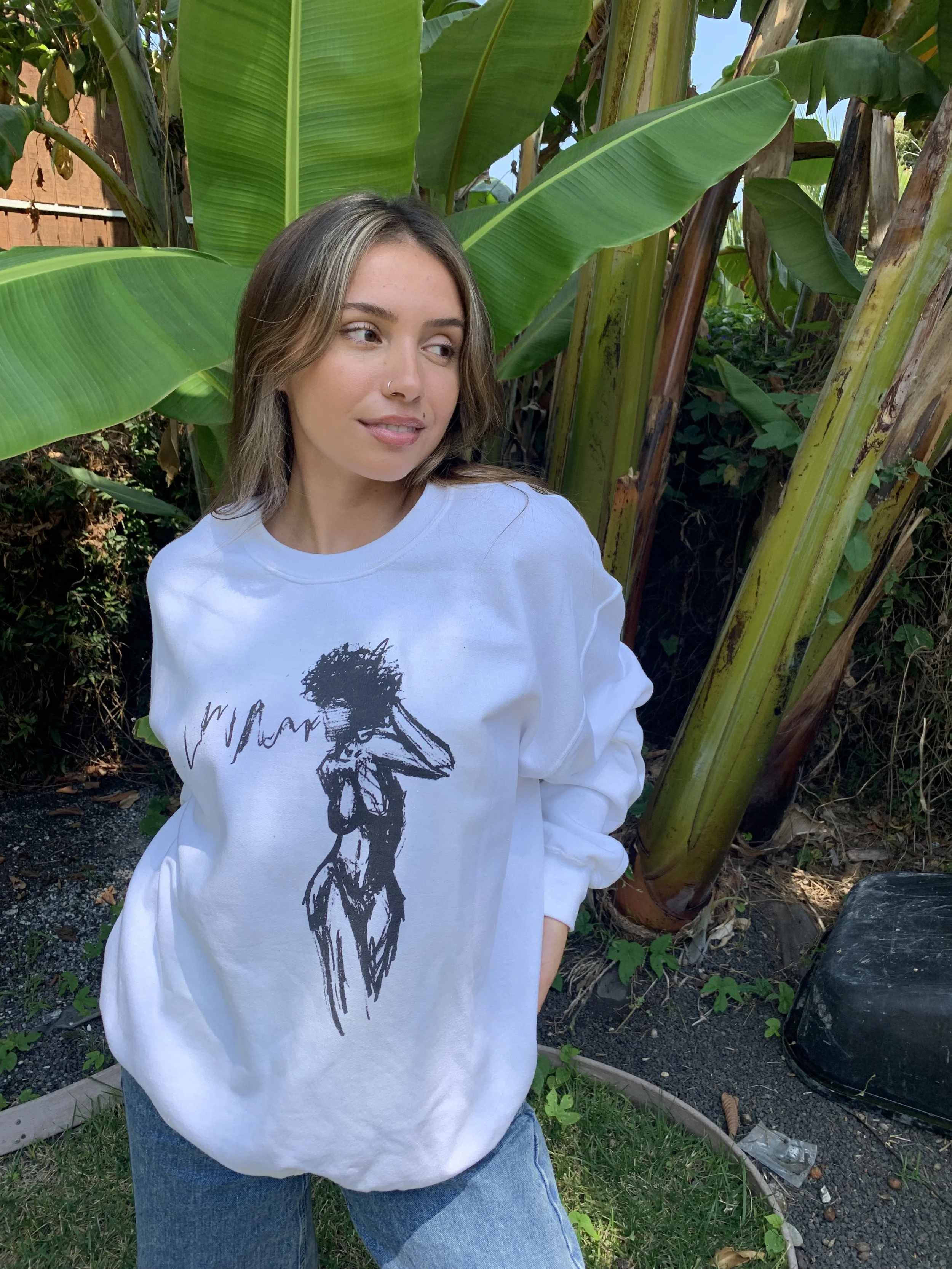 Woman in white sweatshirt with sketch design, standing outdoors in front of large green banana leaves, looking to the side.