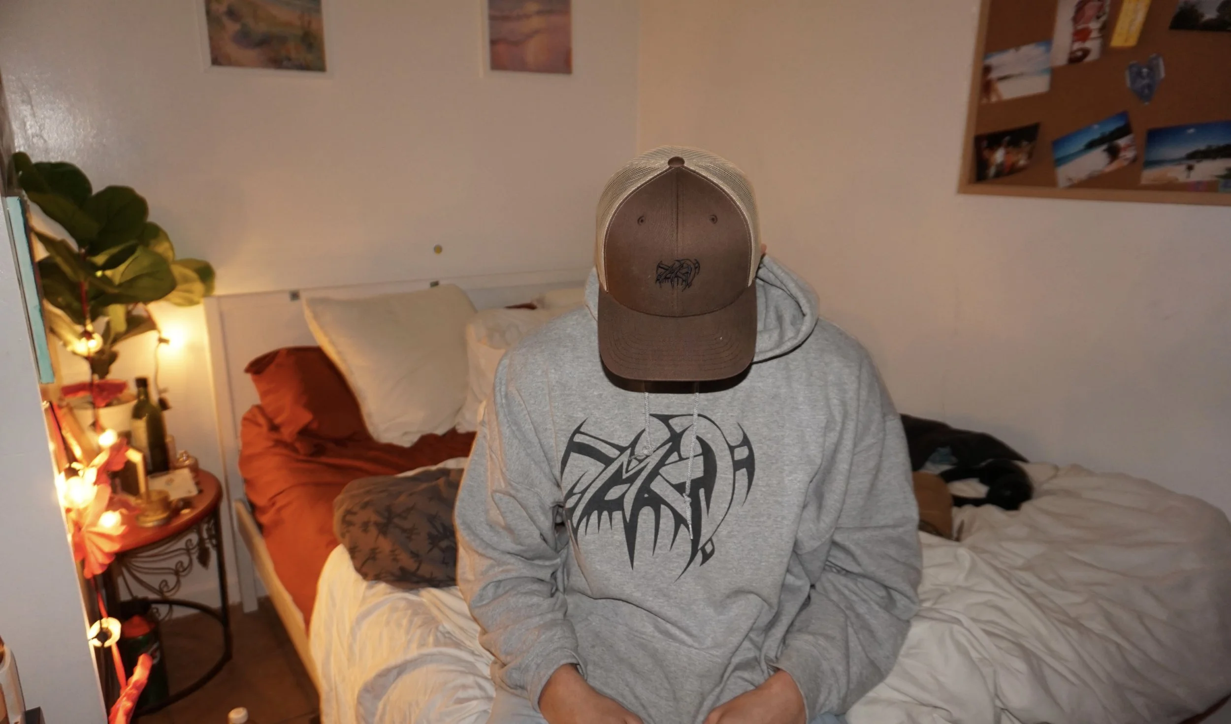 Person wearing a gray hoodie and brown cap sitting on a bed in a cozy room with a small table, plants, and wall decorations.