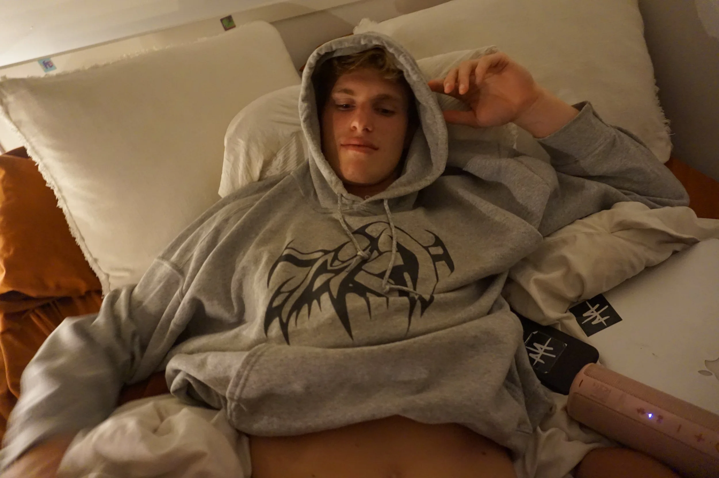 Person lying on a bed wearing a gray hoodie with a tribal design, surrounded by pillows, a laptop, and a speaker.