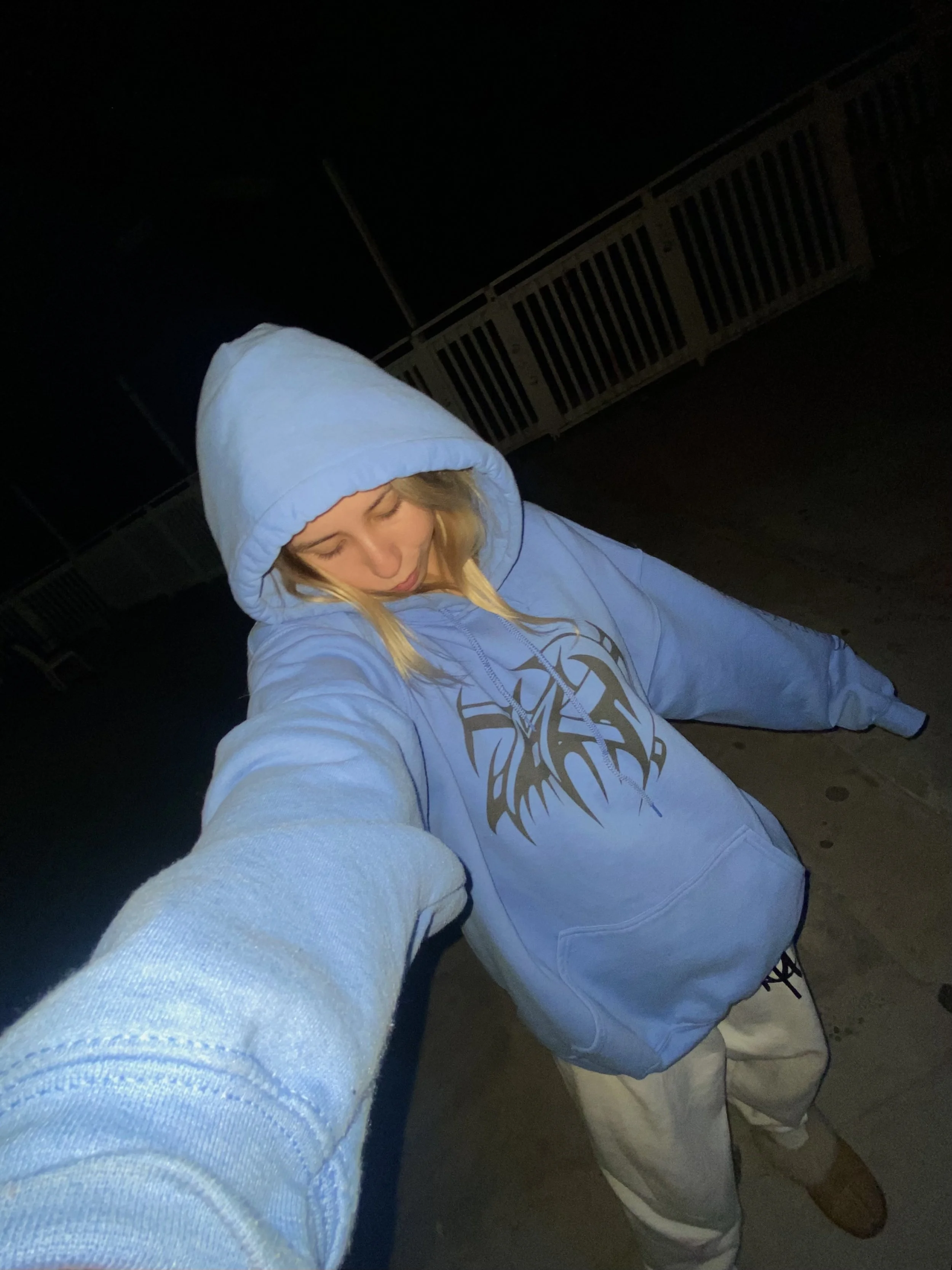 Person wearing a blue hoodie and sweatpants in a dark outdoor area, taking a selfie with head down.