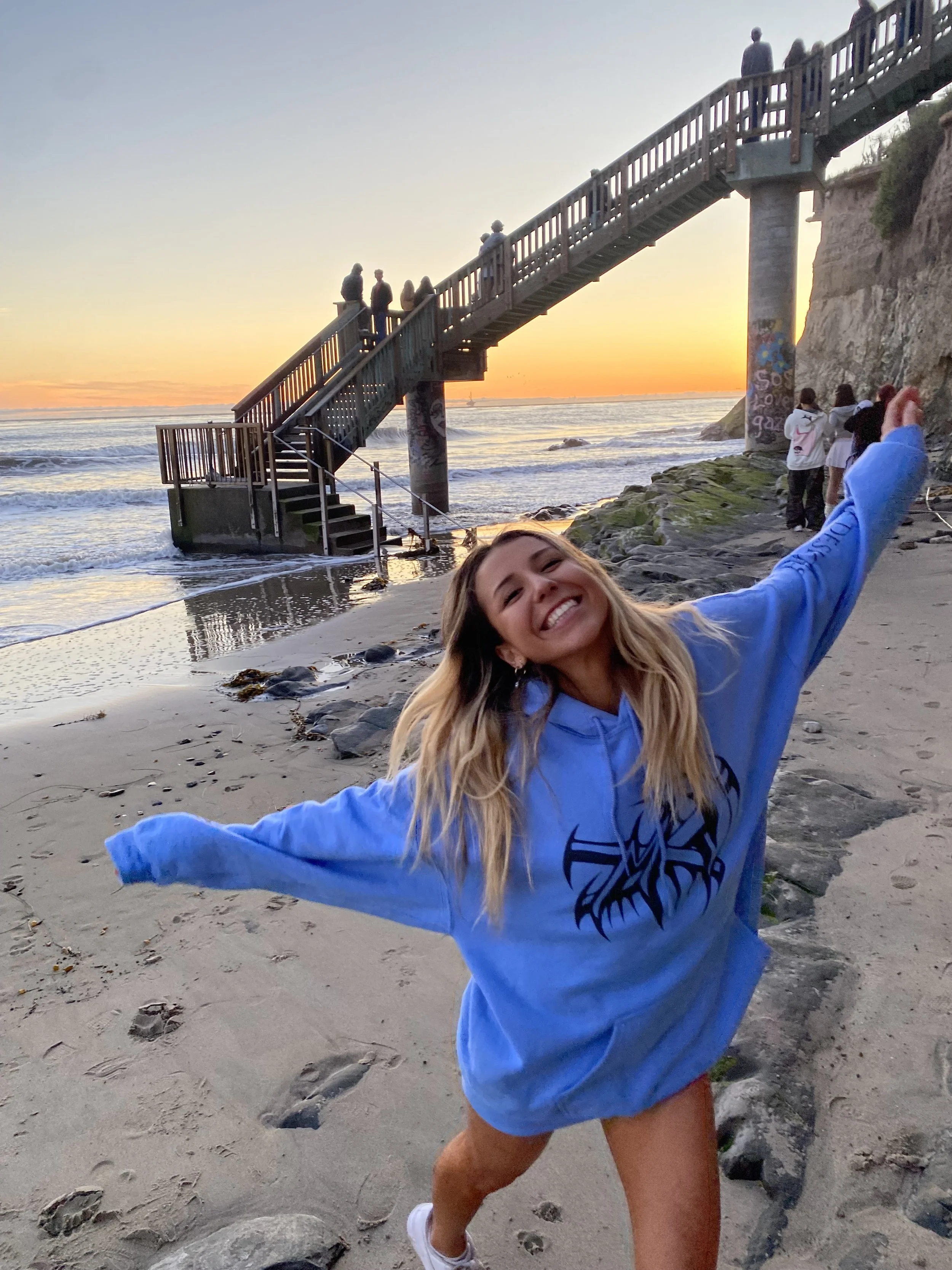Person in a blue hoodie smiling and posing on a beach at sunset, with people on a staircase in the background leading from a cliff down to the shore.