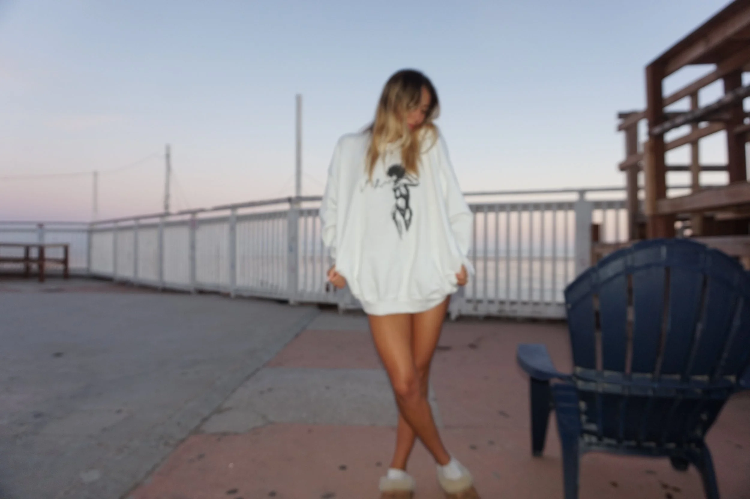 Person in oversized white sweater standing on a beach boardwalk with white railing and Adirondack chair.