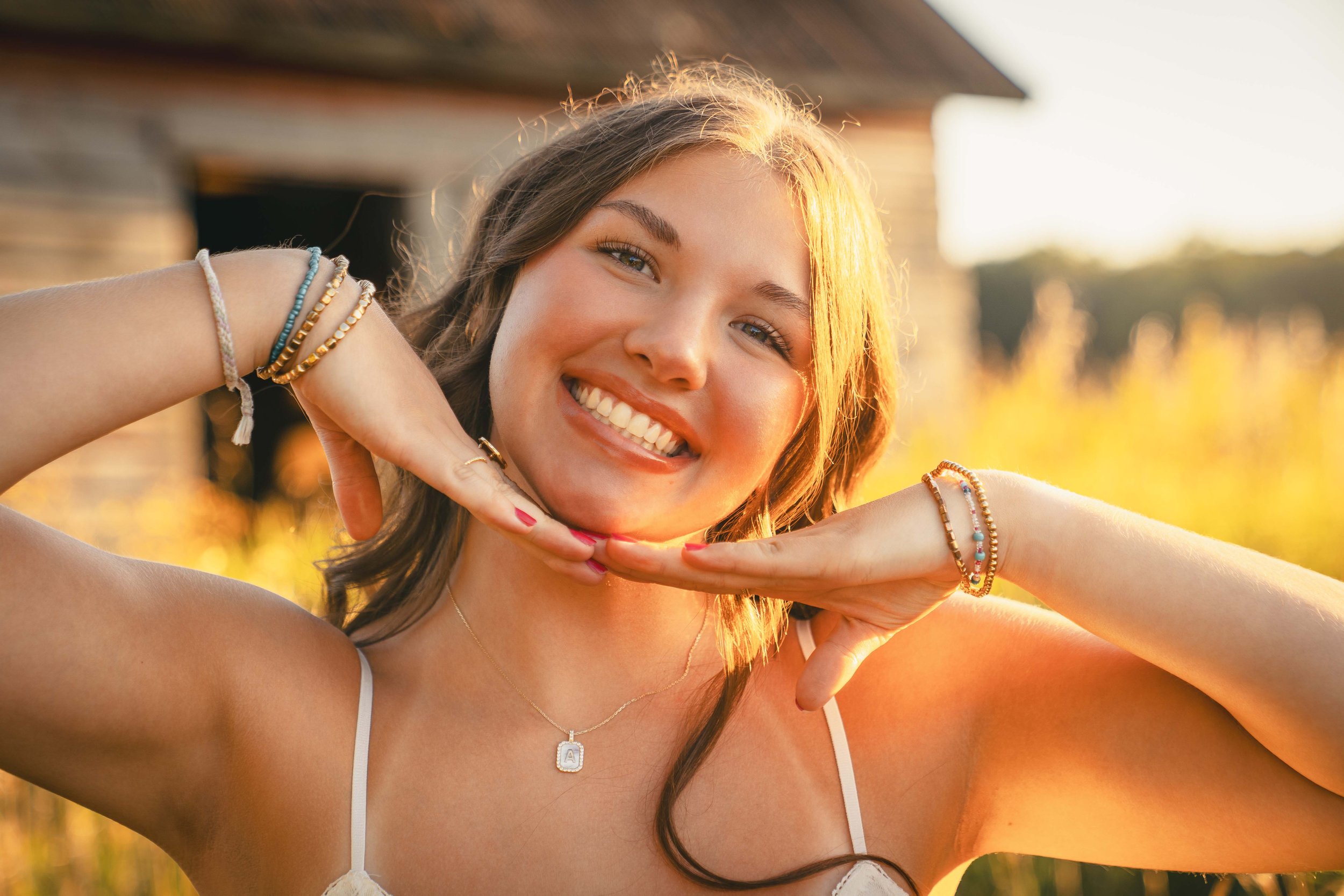 A young woman smiling and posing outdoors during sunset, with her hands under her chin, wearing a white tank top and colorful bracelets.