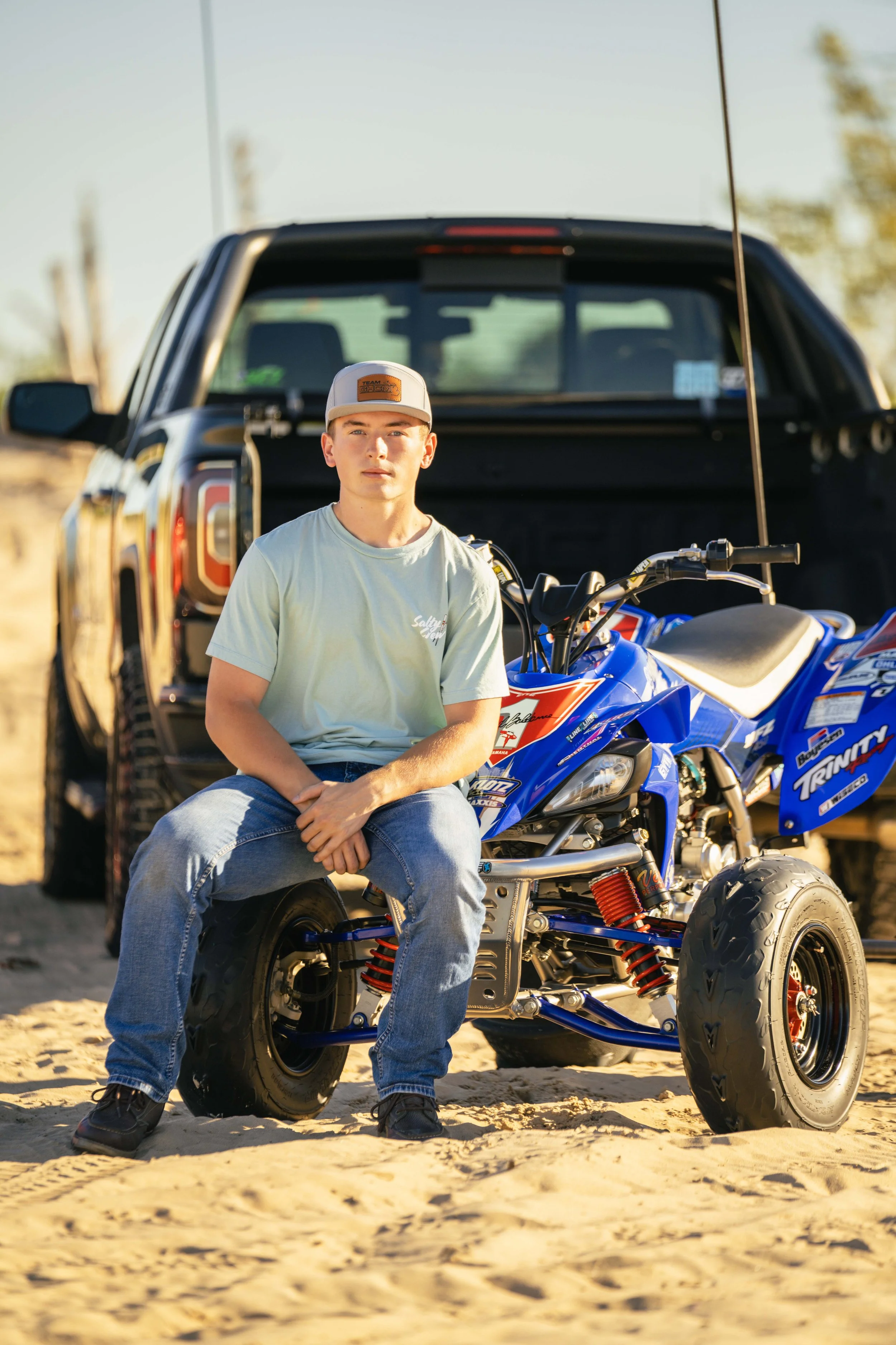 A young man sitting on a small blue all-terrain vehicle (ATV) in front of a black pickup truck on a sandy outdoor setting, with a background of trees and clear sky.