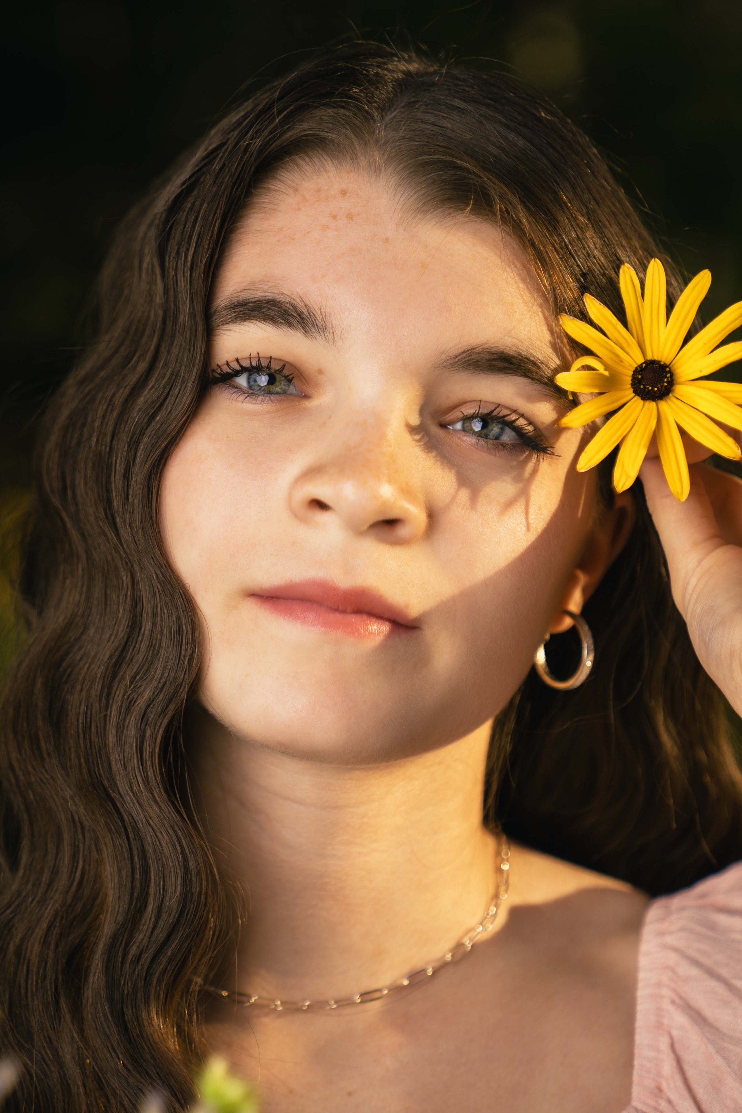 A young woman with wavy brown hair and blue eyes holding a yellow flower near her face