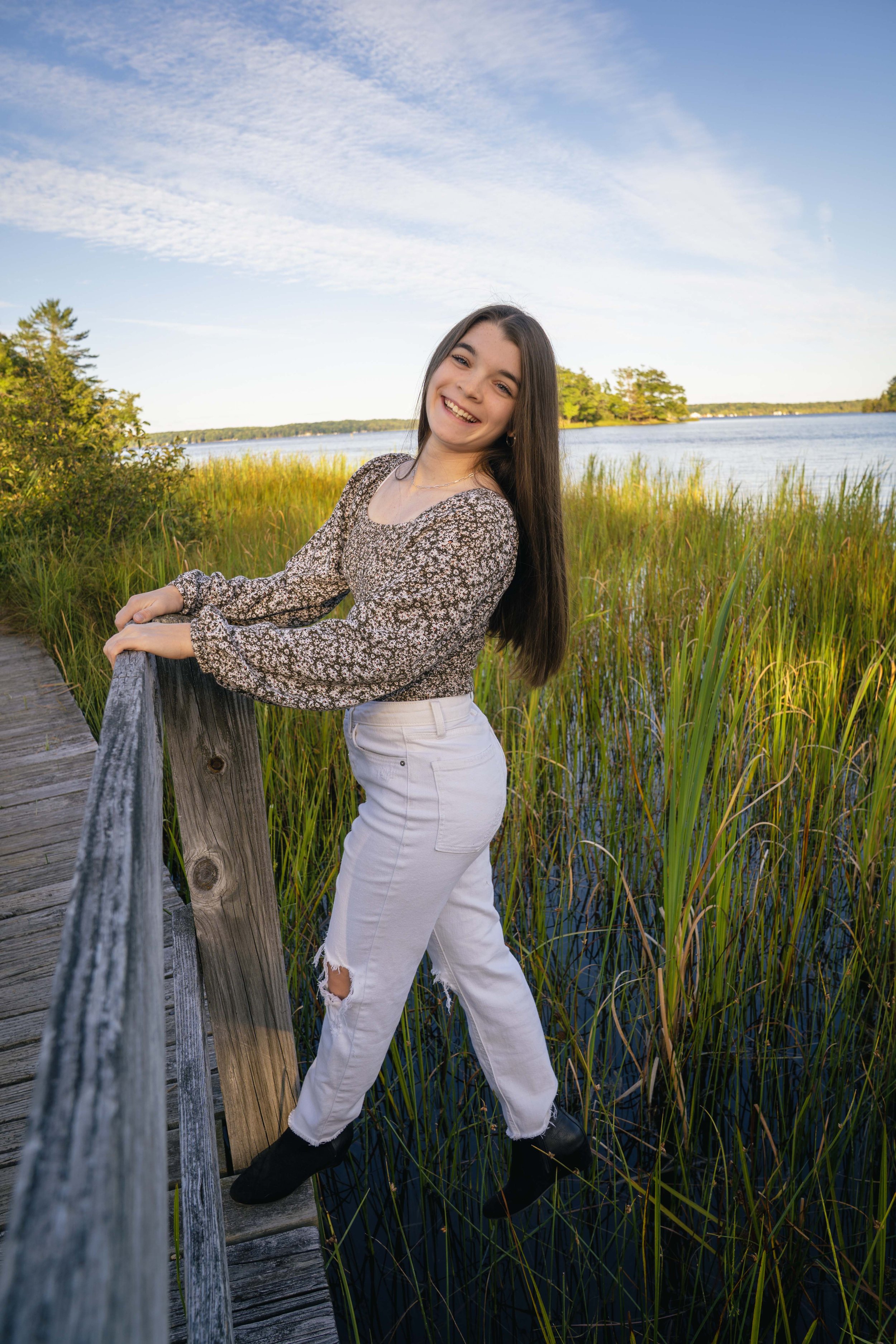 A young woman with long dark hair smiling and leaning on a wooden railing on a boardwalk by a lake during daytime. She is wearing a floral long-sleeve top, white ripped jeans, and black boots, with tall grass and trees in the background.