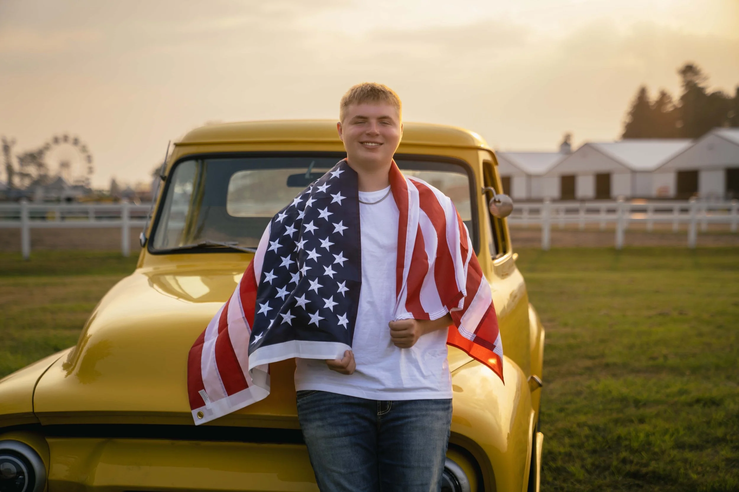 A young man is standing in front of a yellow vehicle at sunset, draped in an American flag, smiling.