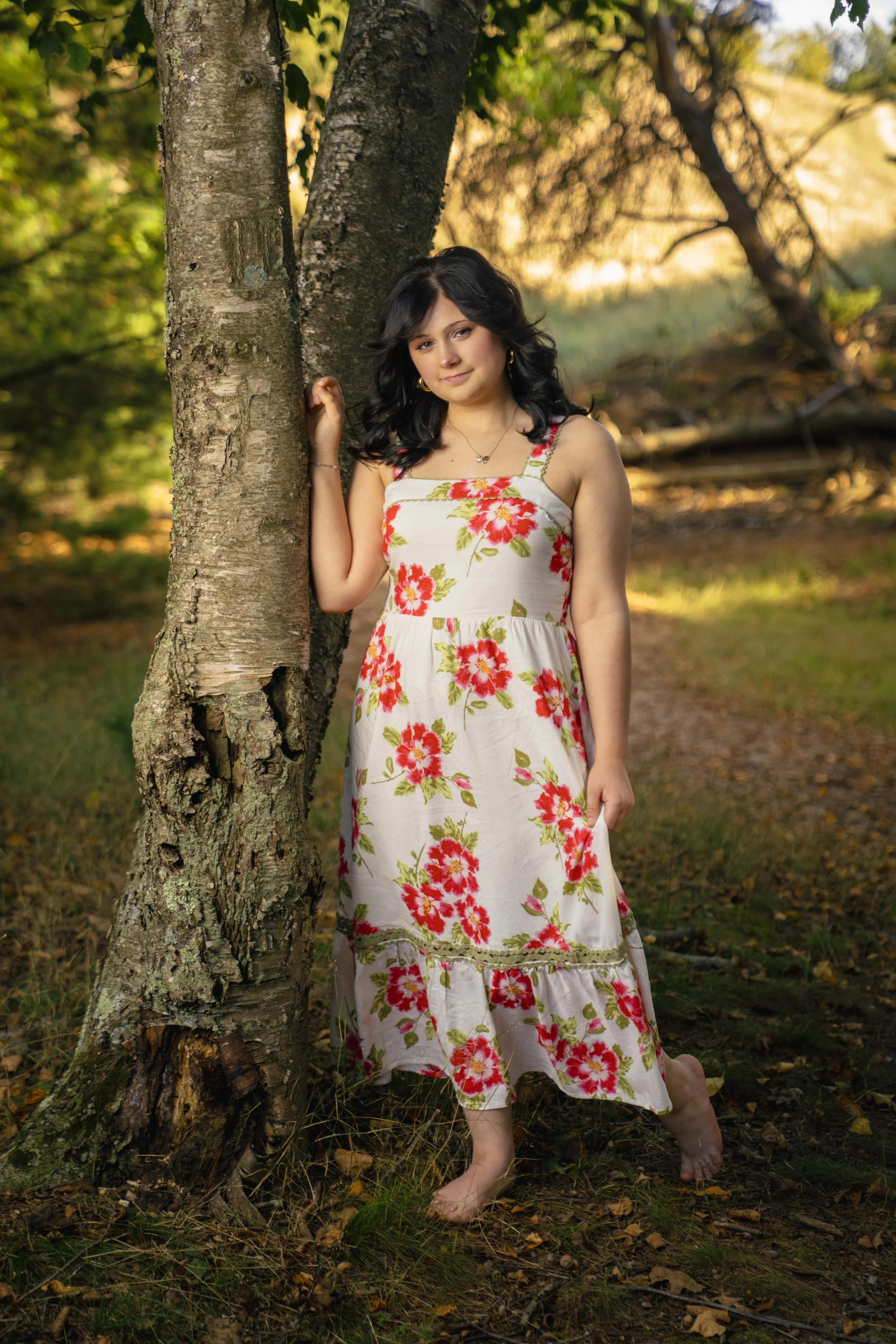 A young woman with dark hair in a floral dress standing barefoot outdoors near a tree with falling leaves and a sunset in the background.