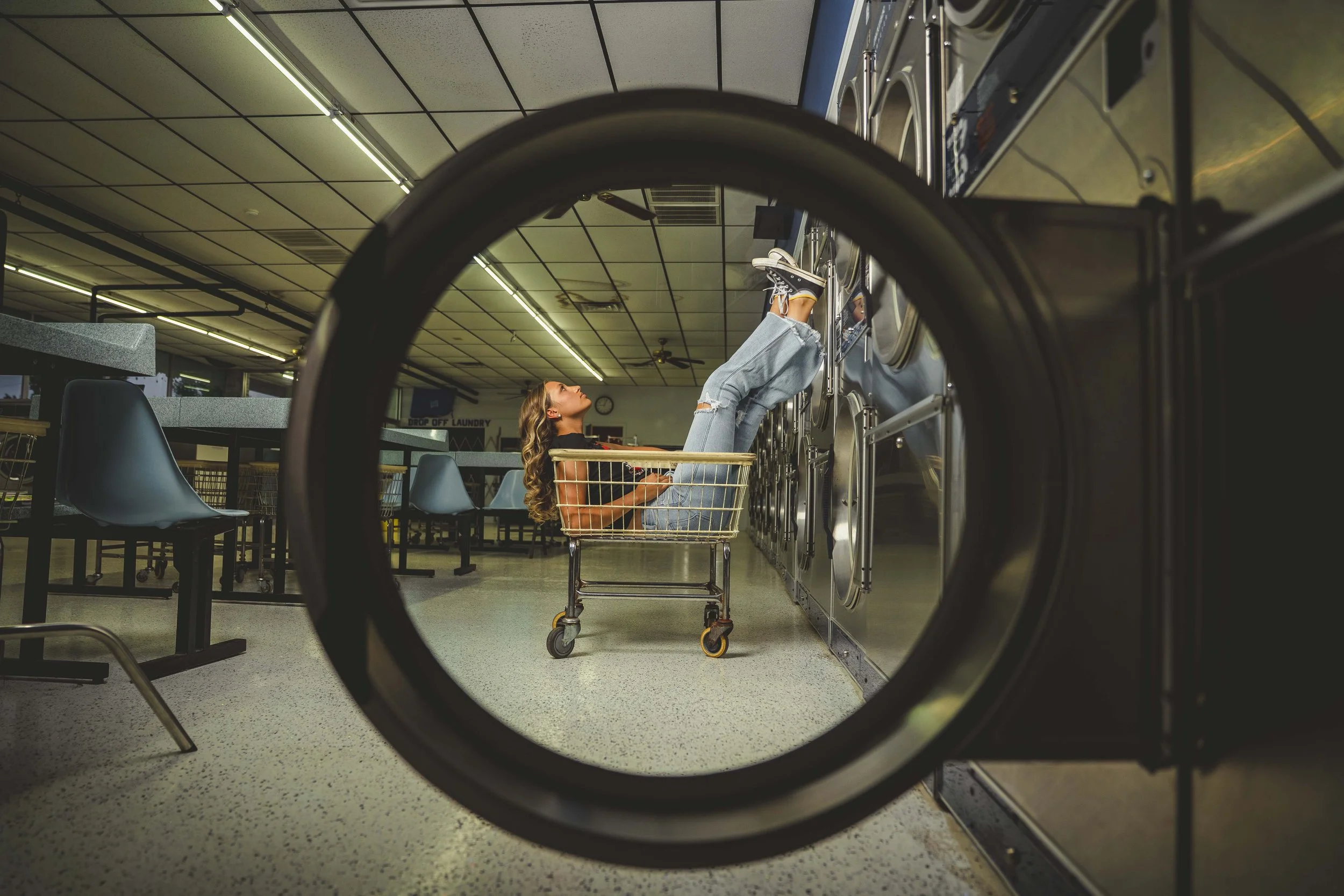A woman lying in a laundry cart with her legs raised and feet on a row of washing machines in a laundromat.