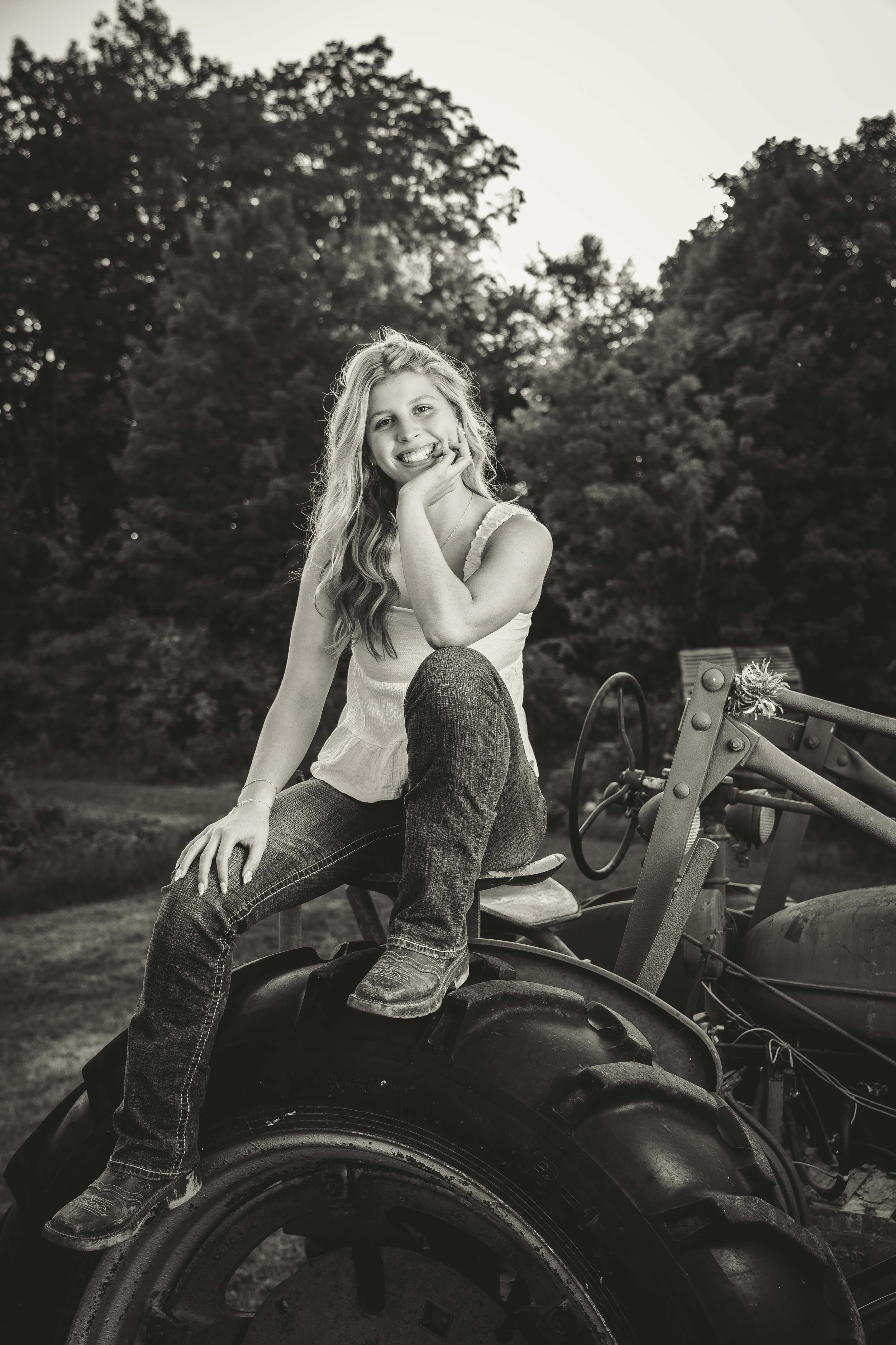 A young woman sitting on a tractor outdoors, smiling and resting her chin on her hand, with trees in the background.