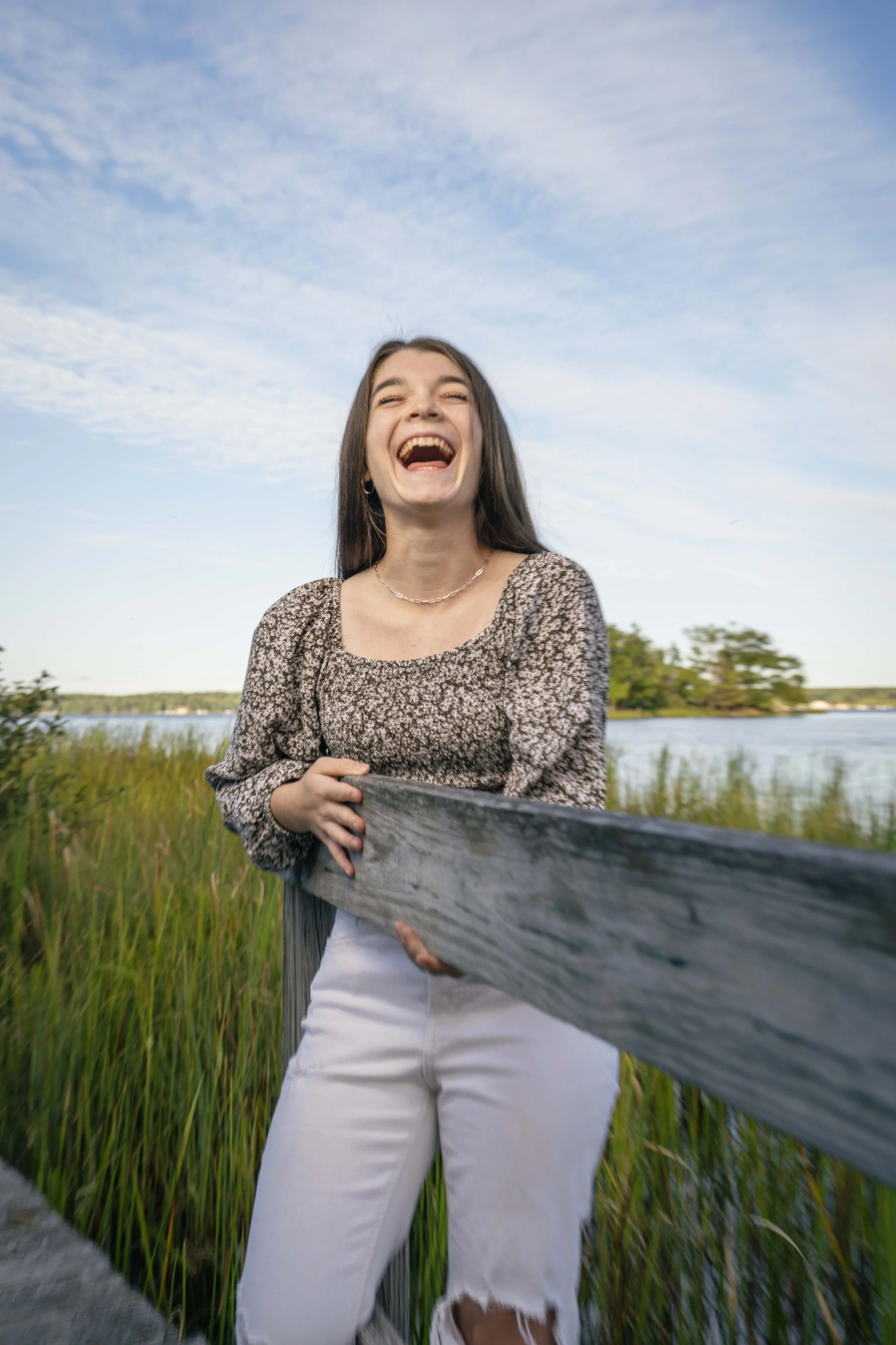 Young woman with long brown hair, laughing and holding onto a wooden railing outdoors near a body of water with tall grasses and trees in the background, under a partly cloudy sky.