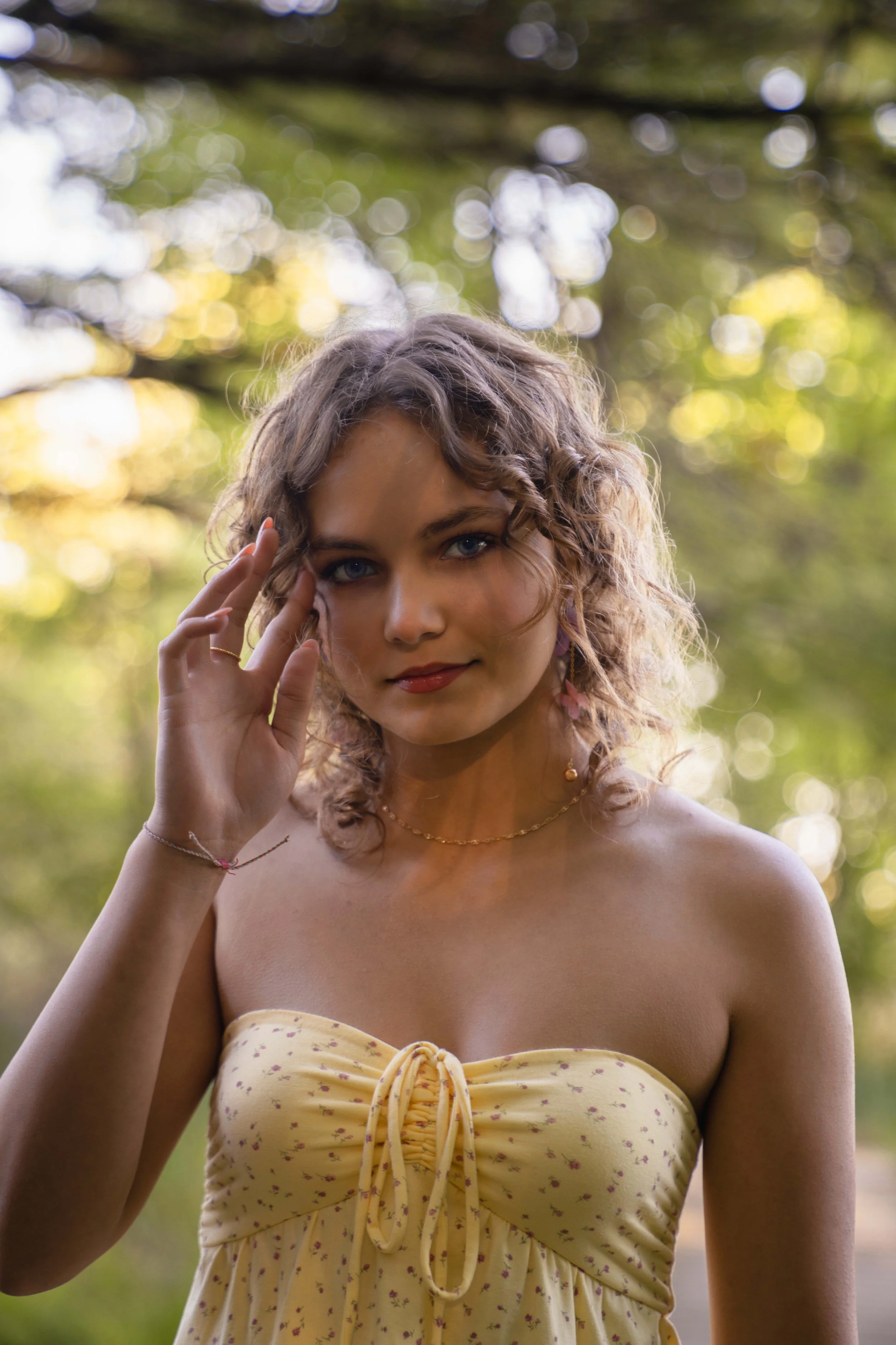 A young woman with curly hair and blue eyes, wearing a yellow strapless dress with a floral pattern, standing outdoors with trees and sunlight in the background.