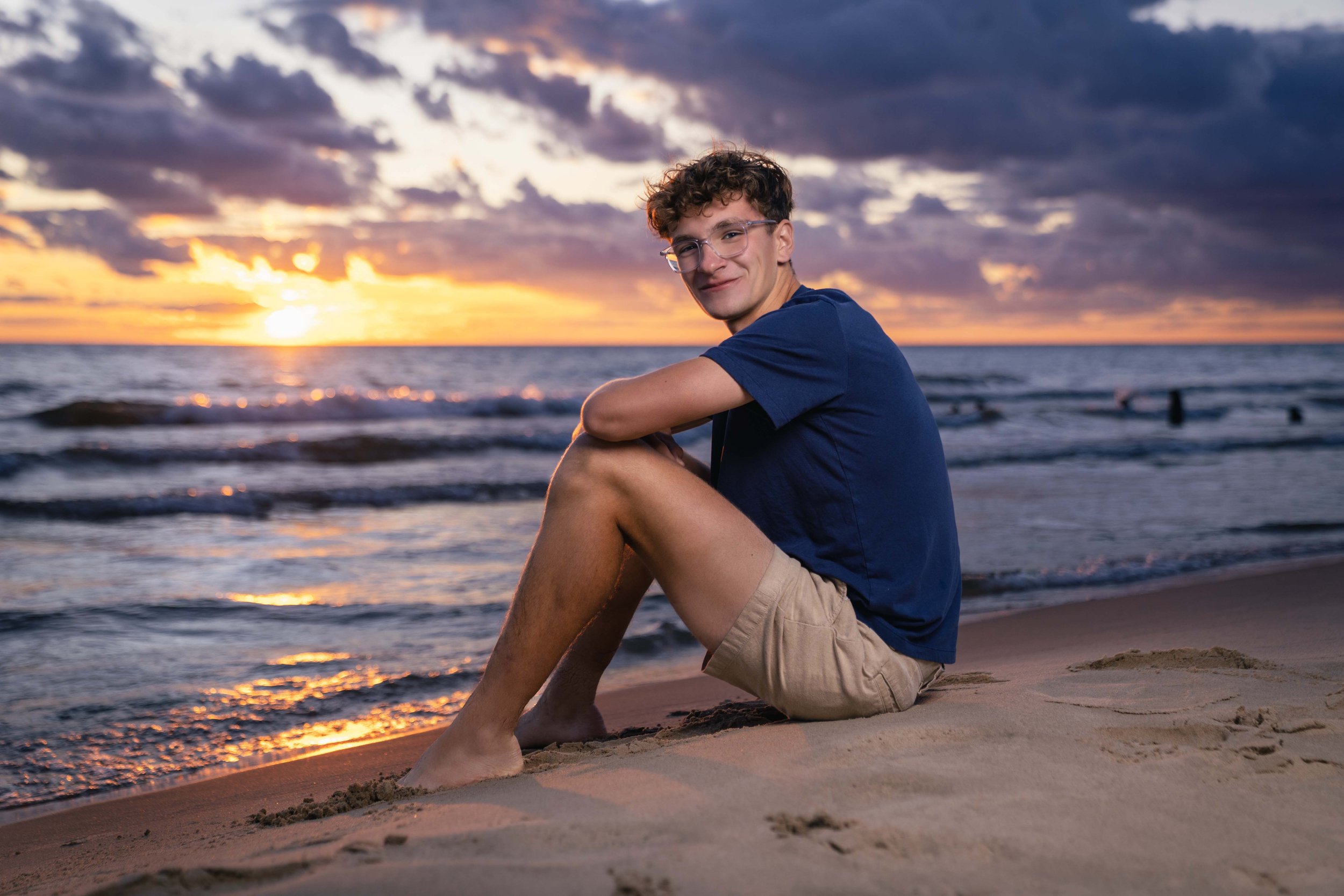 A teenage boy sitting on the sandy beach at sunset, with the ocean in the background, wearing glasses, a blue t-shirt, and beige shorts, smiling at the camera.