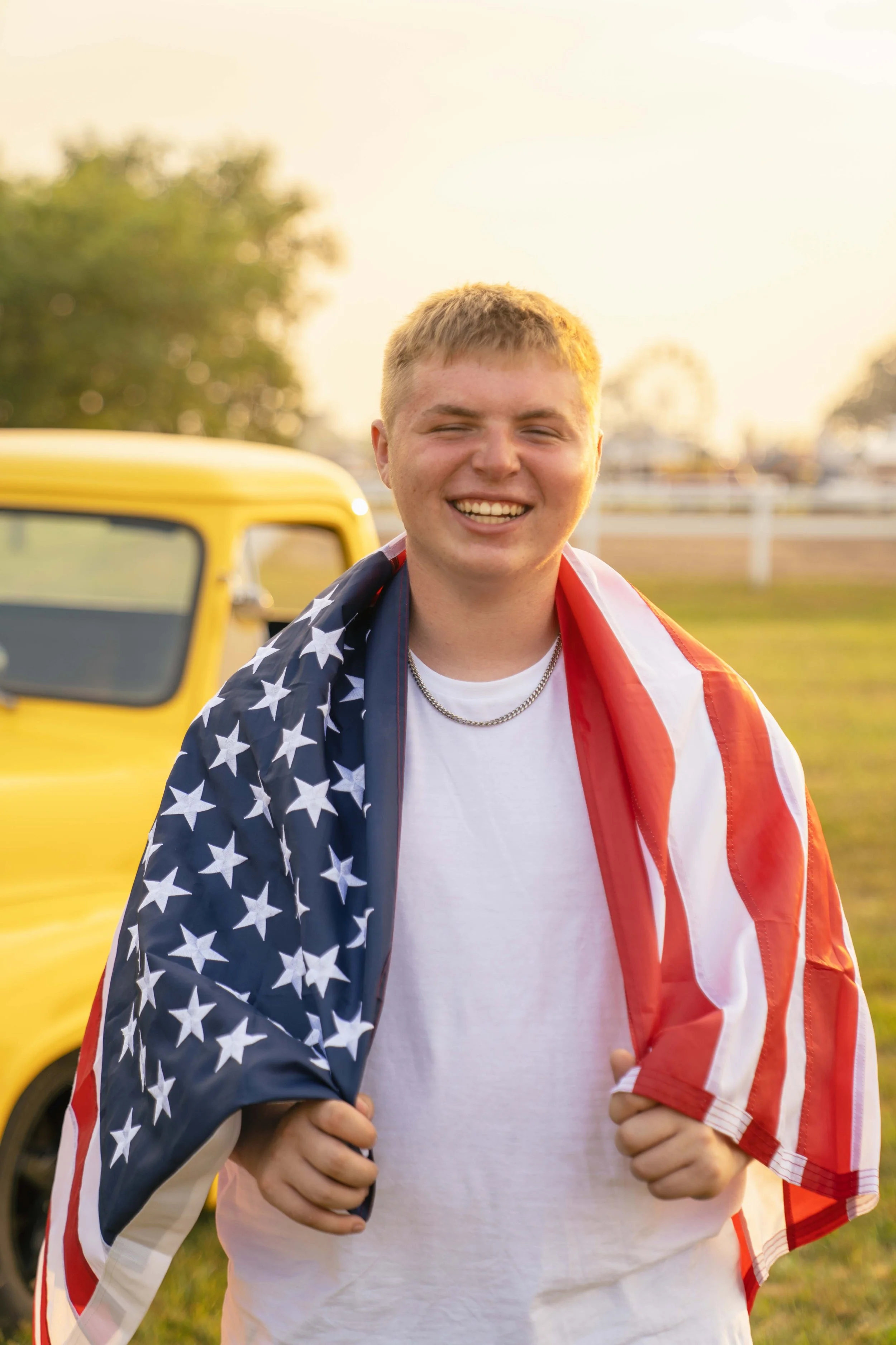 A young man smiling outdoors, draped in an American flag, standing in front of a yellow vehicle at sunset.