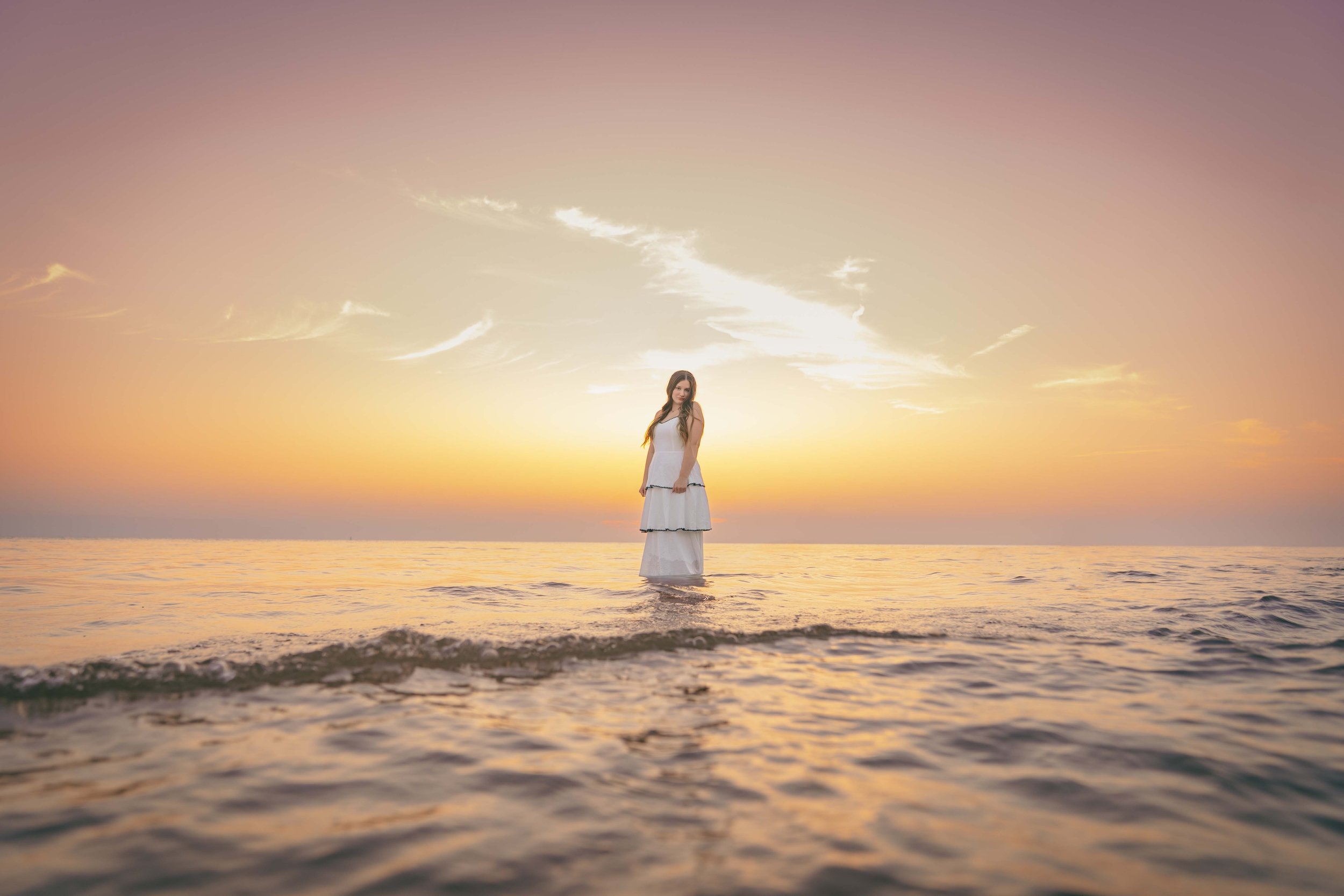 Woman in white dress standing in shallow water during sunset over the ocean.