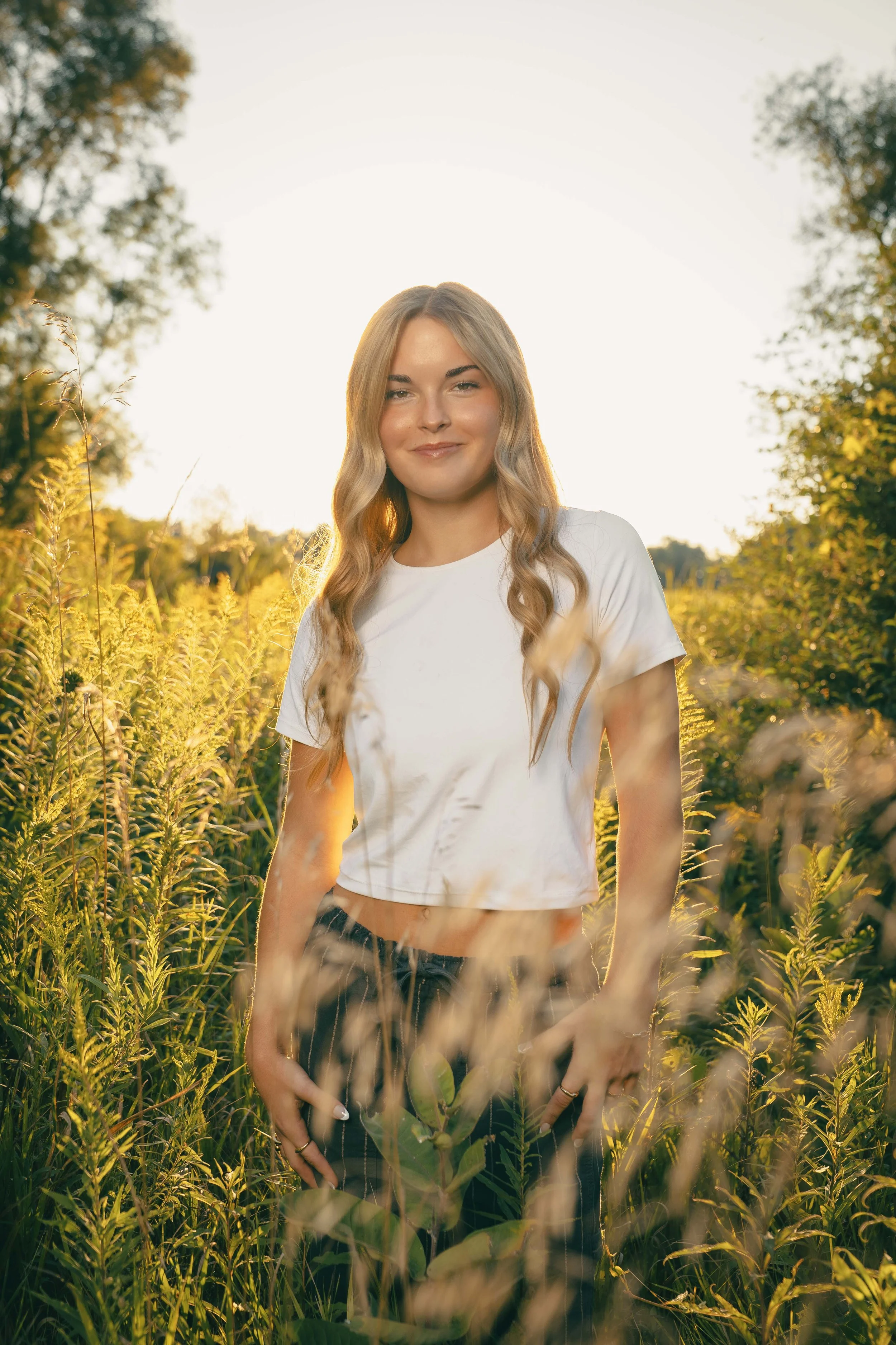 Young woman with blonde wavy hair smiling in a sunlit field of tall grass and plants during sunset.